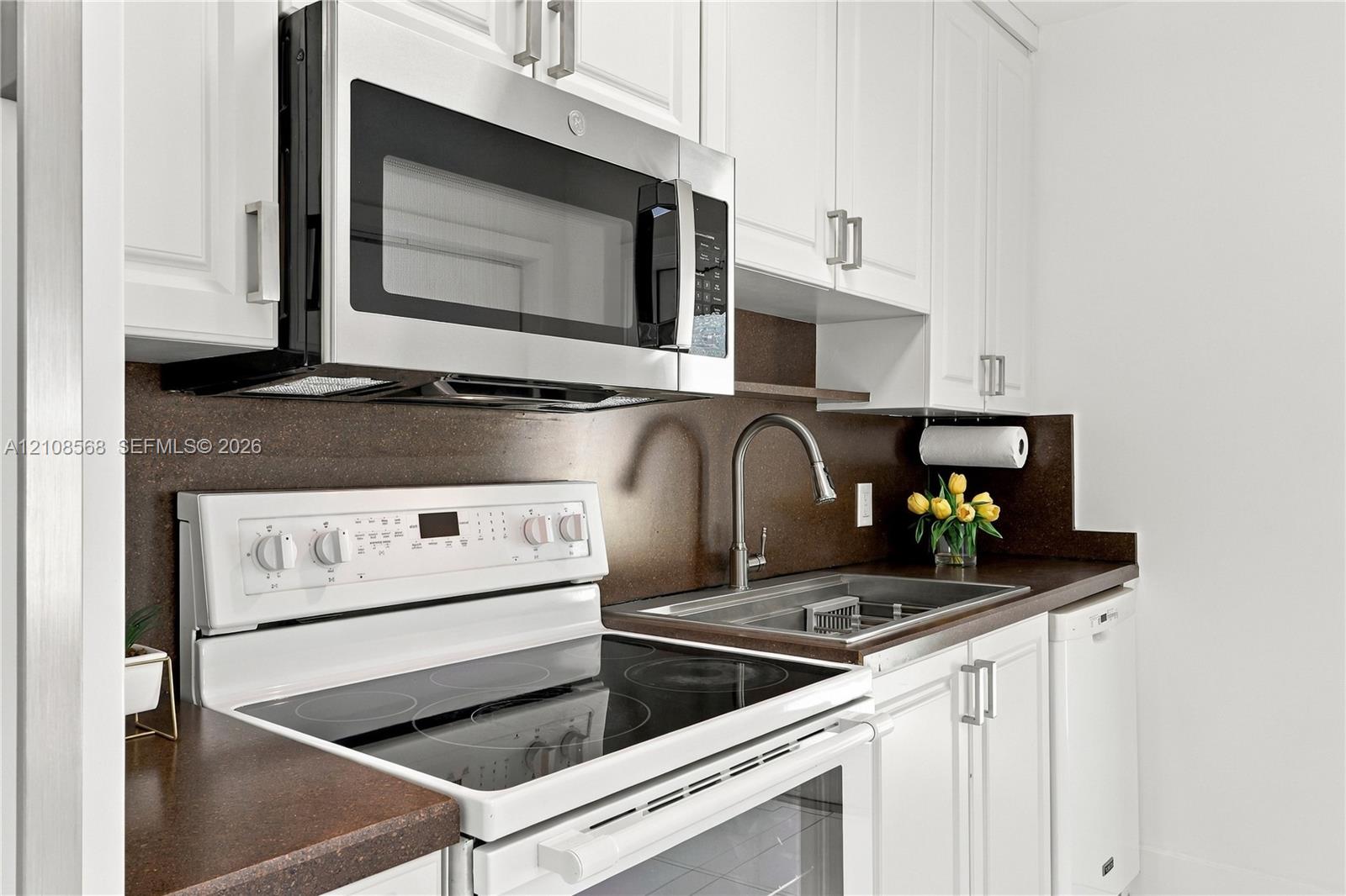 White shaker-style cabinetry, stainless steel over-the-range microwave, glass cooktop range, integrated stainless steel sink with high-arc faucet, and solid-surface countertops