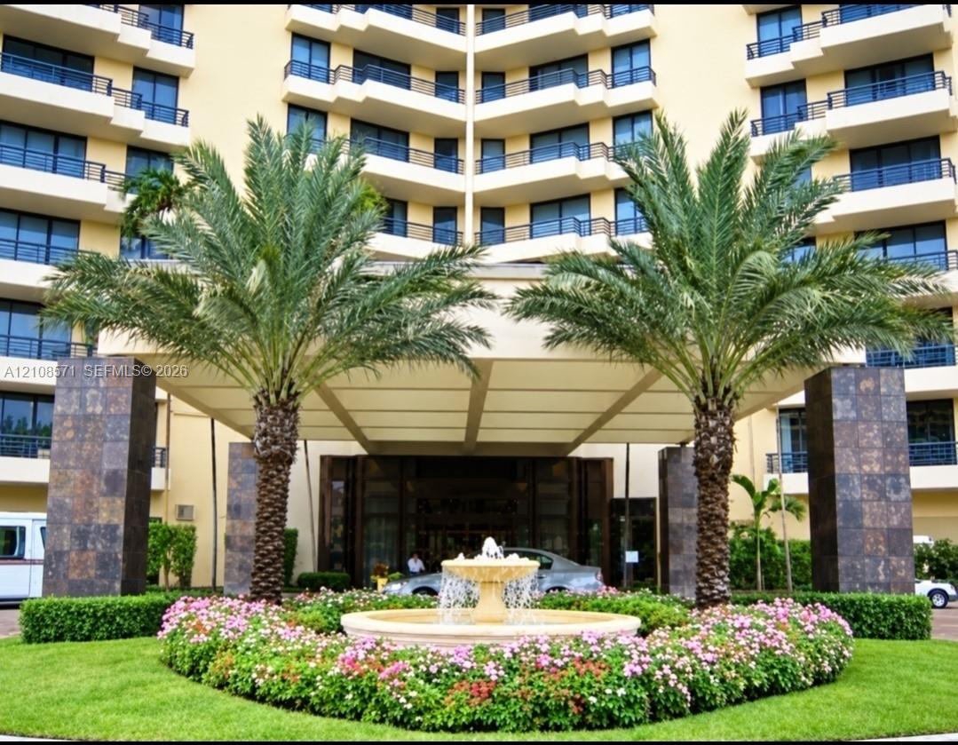 Multi-story building facade featuring a covered porte-cochre, decorative central stone fountain, mature palm trees, and manicured landscaping with flowering perennials