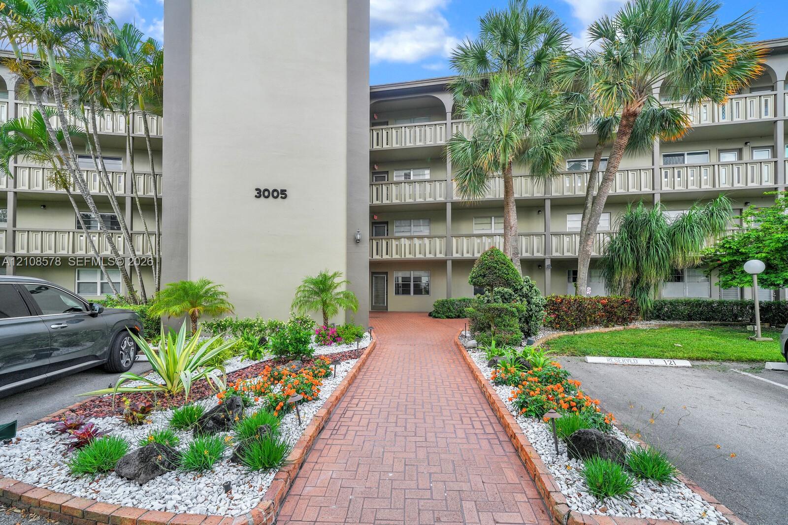 Multi-story building exterior featuring covered balconies, brick-paver walkway, and mature tropical landscaping