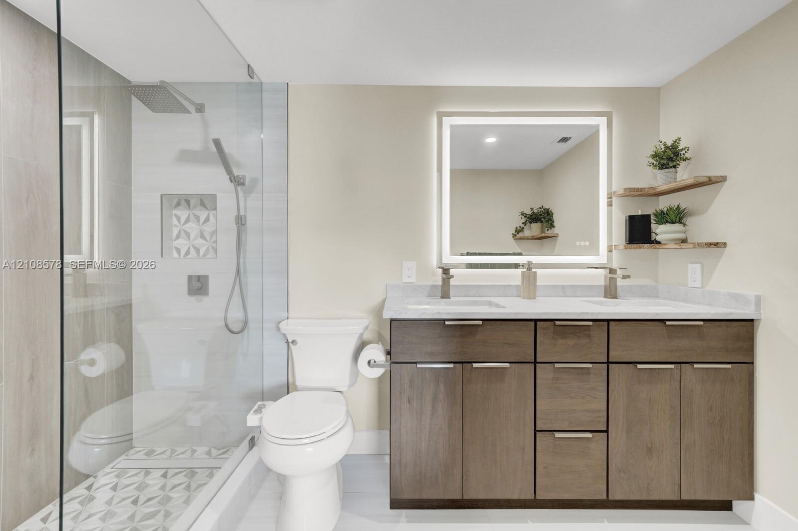 Bathroom featuring a dual-sink vanity with wood-finish cabinetry, a backlit mirror, a glass-enclosed shower with patterned tile inlay, and floating wood-finished shelving