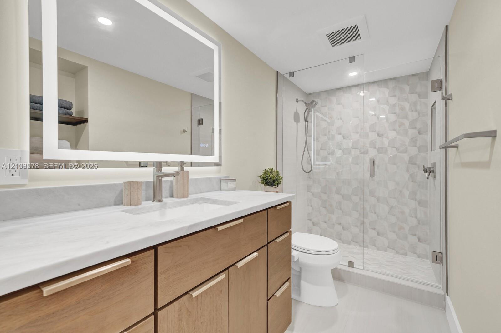 Wood-finish vanity with a stone countertop, illuminated rectangular mirror, and a glass-enclosed walk-in shower featuring geometric tile accents