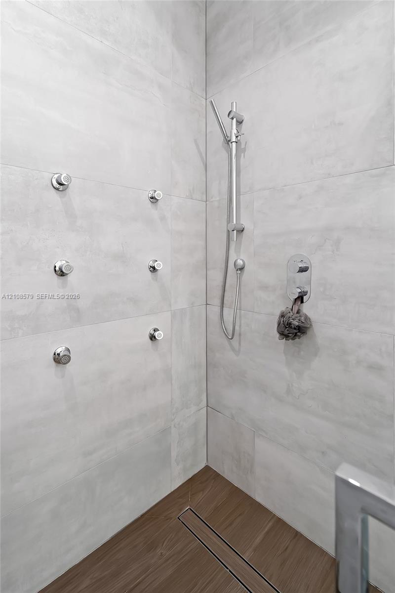 Walk-in shower featuring large-format light grey wall tile, six integrated body sprays, a handheld showerhead on a slide bar, and wood-finish flooring with a linear drain