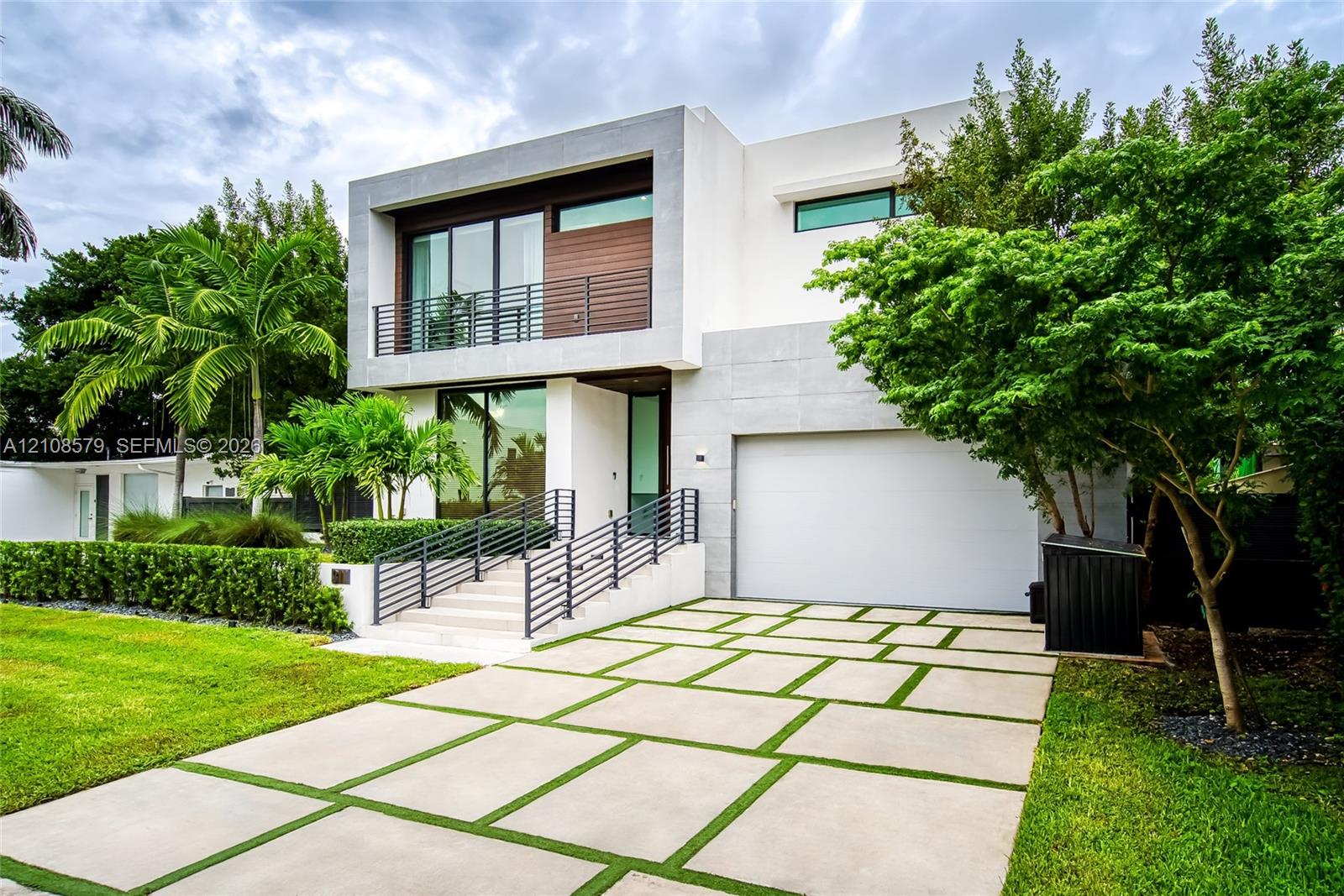 Modern two-story architectural exterior featuring smooth-finish stucco walls, natural wood-paneled accents, a linear upper-level balcony with metal railings, and a geometric concrete paver driveway with integrated turf strips