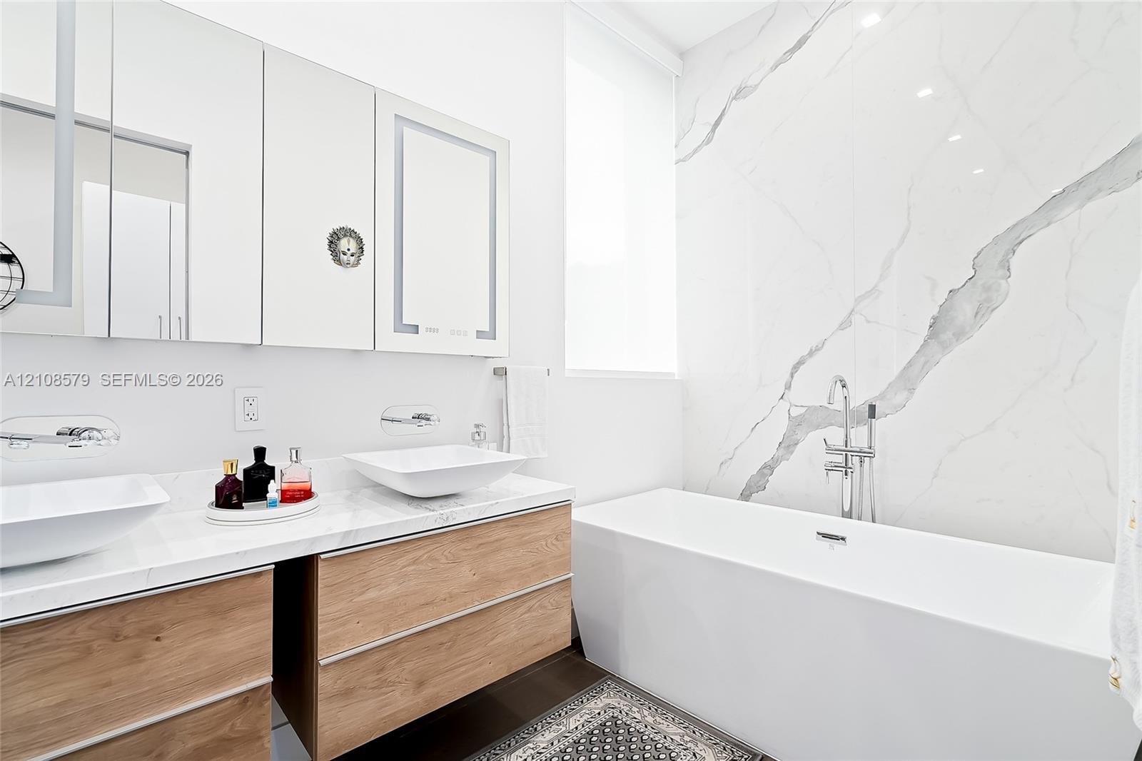 Double floating vanity with integrated rectangular vessel sinks and wood-finish cabinetry, complemented by a freestanding soaking tub, large-format stone-patterned wall tile, and built-in medicine cabinets with recessed LED lighting