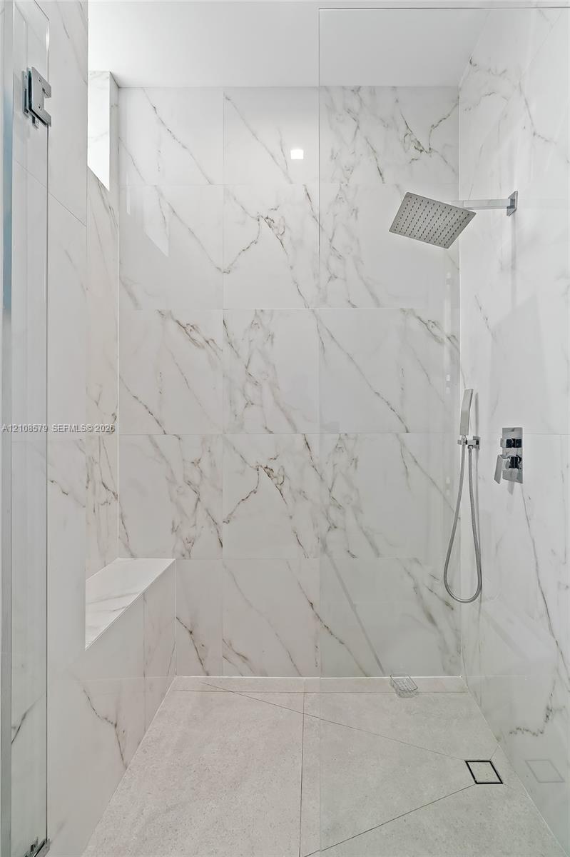 Walk-in shower featuring floor-to-ceiling veined stone-finish tile, glass enclosure, built-in tiled bench, rainfall showerhead, and separate handheld sprayer