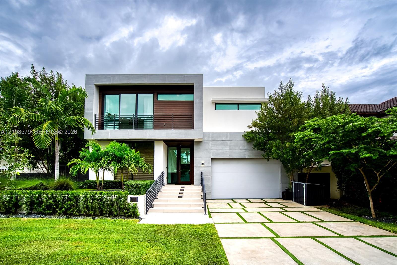 Contemporary two-story exterior featuring a stone-clad facade, wood-panel siding, large-format paver driveway with turf accents, second-story balcony with metal railings, and wide concrete steps leading to the main entrance