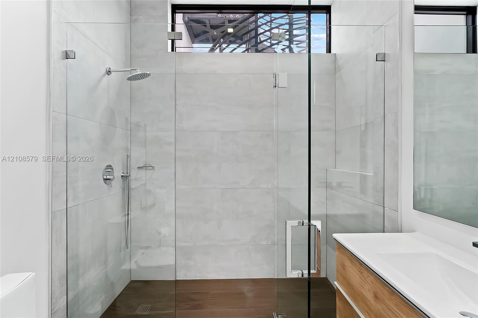 Walk-in shower featuring large-format stone-finish wall tile, frameless glass enclosure, chrome rainfall showerhead, and wood-finish cabinetry with an integrated white sink