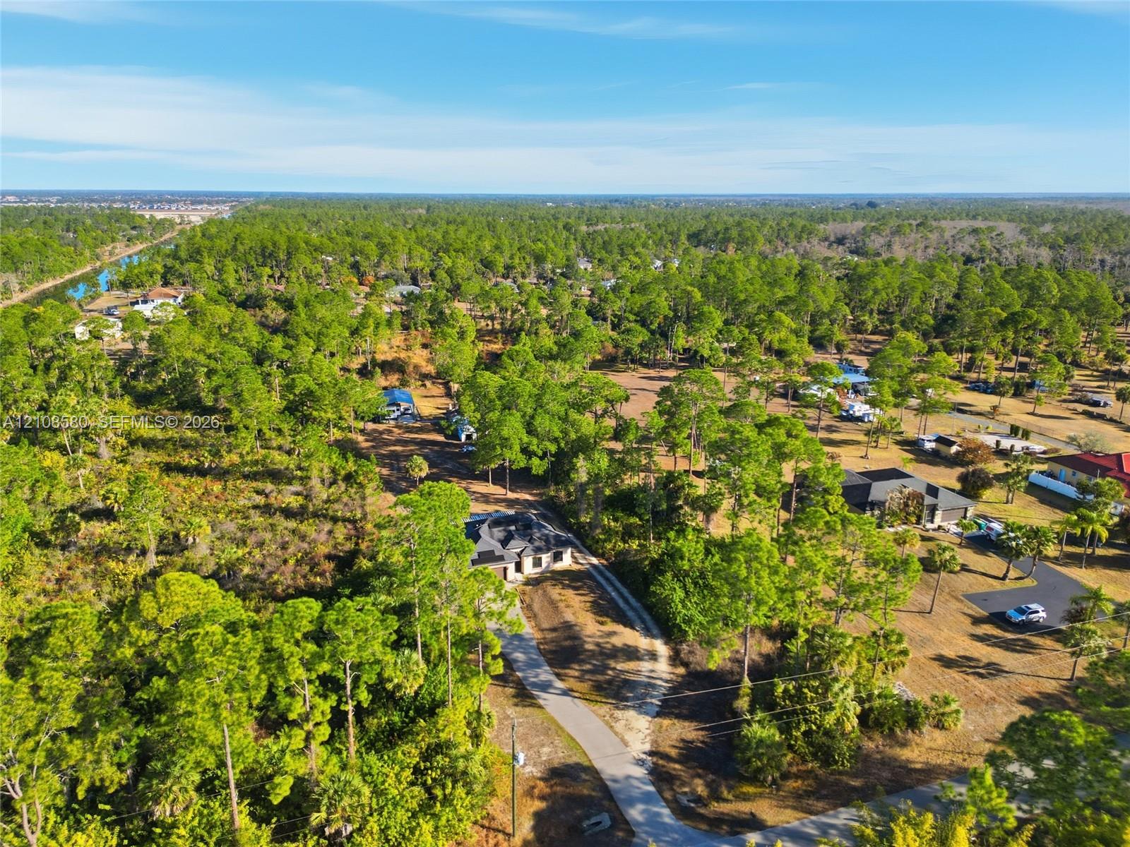 Aerial perspective of a multi-structure property featuring a pitched-roof dwelling, a paved driveway, and expansive natural acreage with dense mature pine vegetation