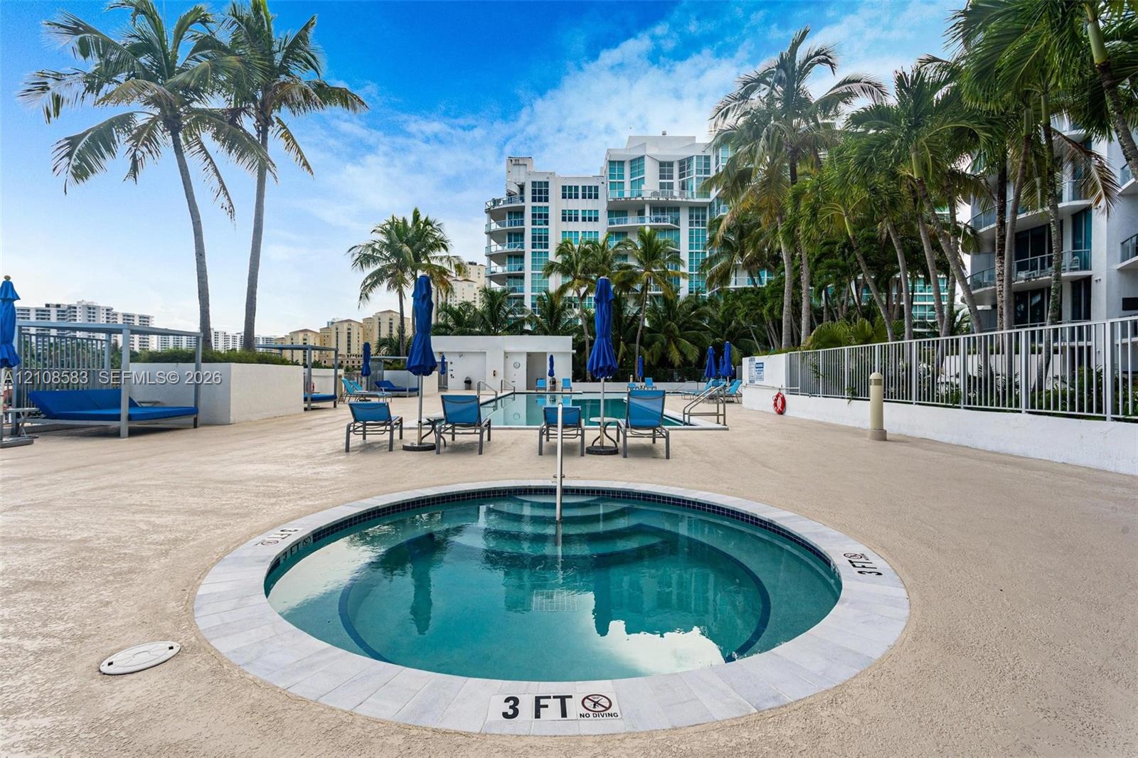 In-ground circular spa with stone coping, concrete decking, mature palm trees, perimeter metal fencing, and an expansive outdoor swimming pool
