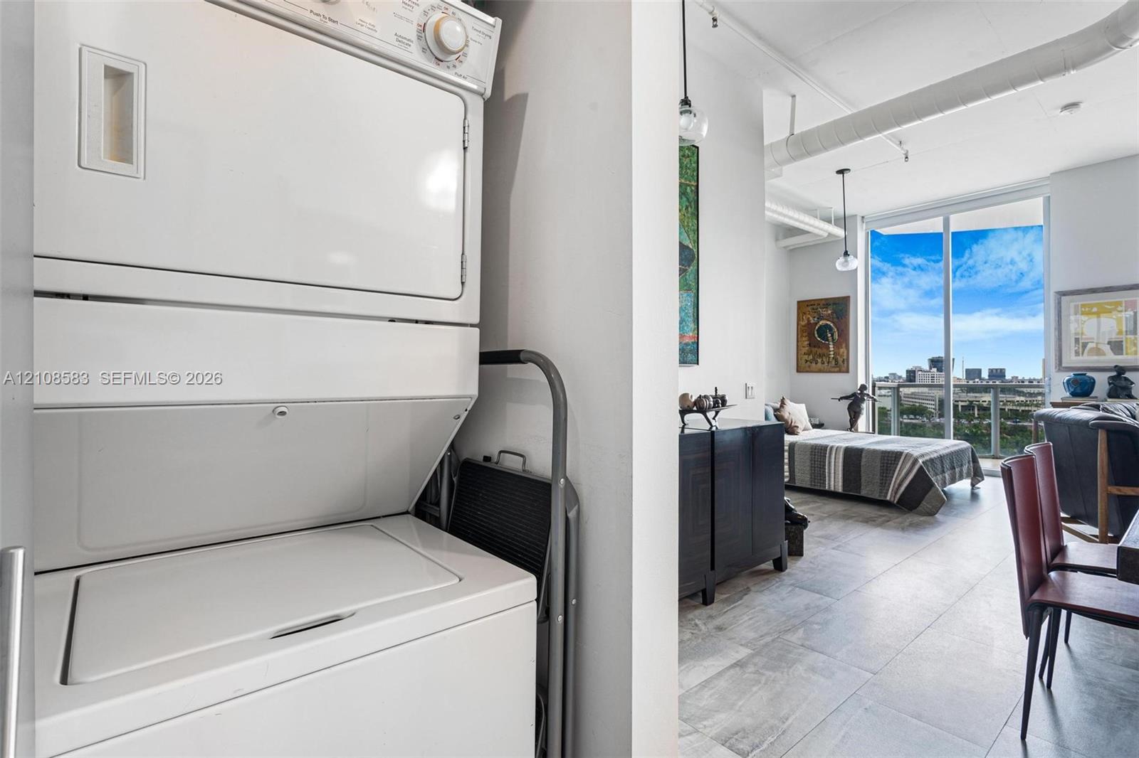 Stacked laundry center featuring white appliance finish, ceramic tile flooring, exposed ductwork, and floor-to-ceiling glass sliding doors