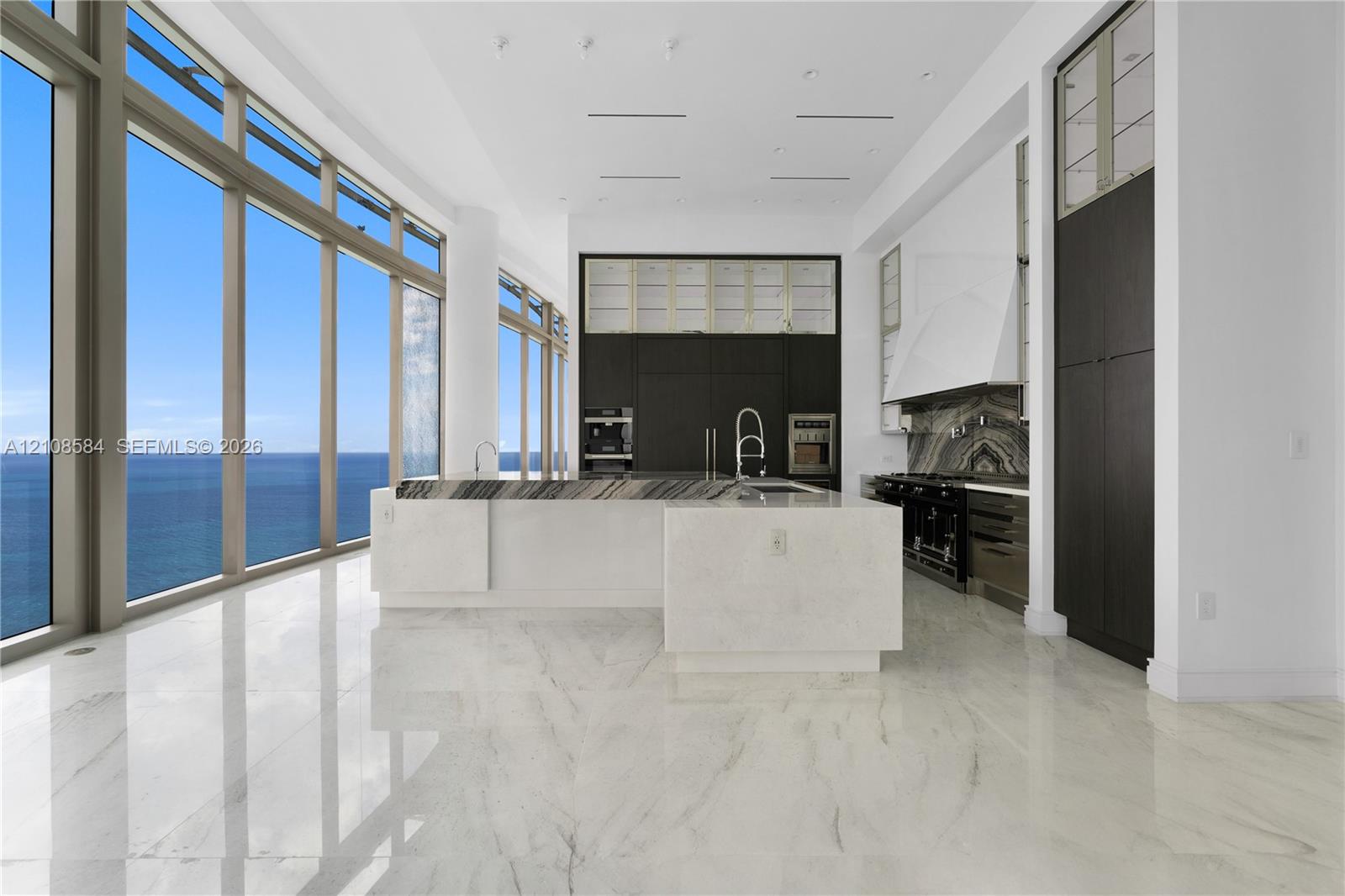 Floor-to-ceiling windows with ocean scenery, expansive center island with stone countertop, custom floor-to-ceiling dark cabinetry, high-gloss stone flooring, and professional-grade range with decorative stone backsplash