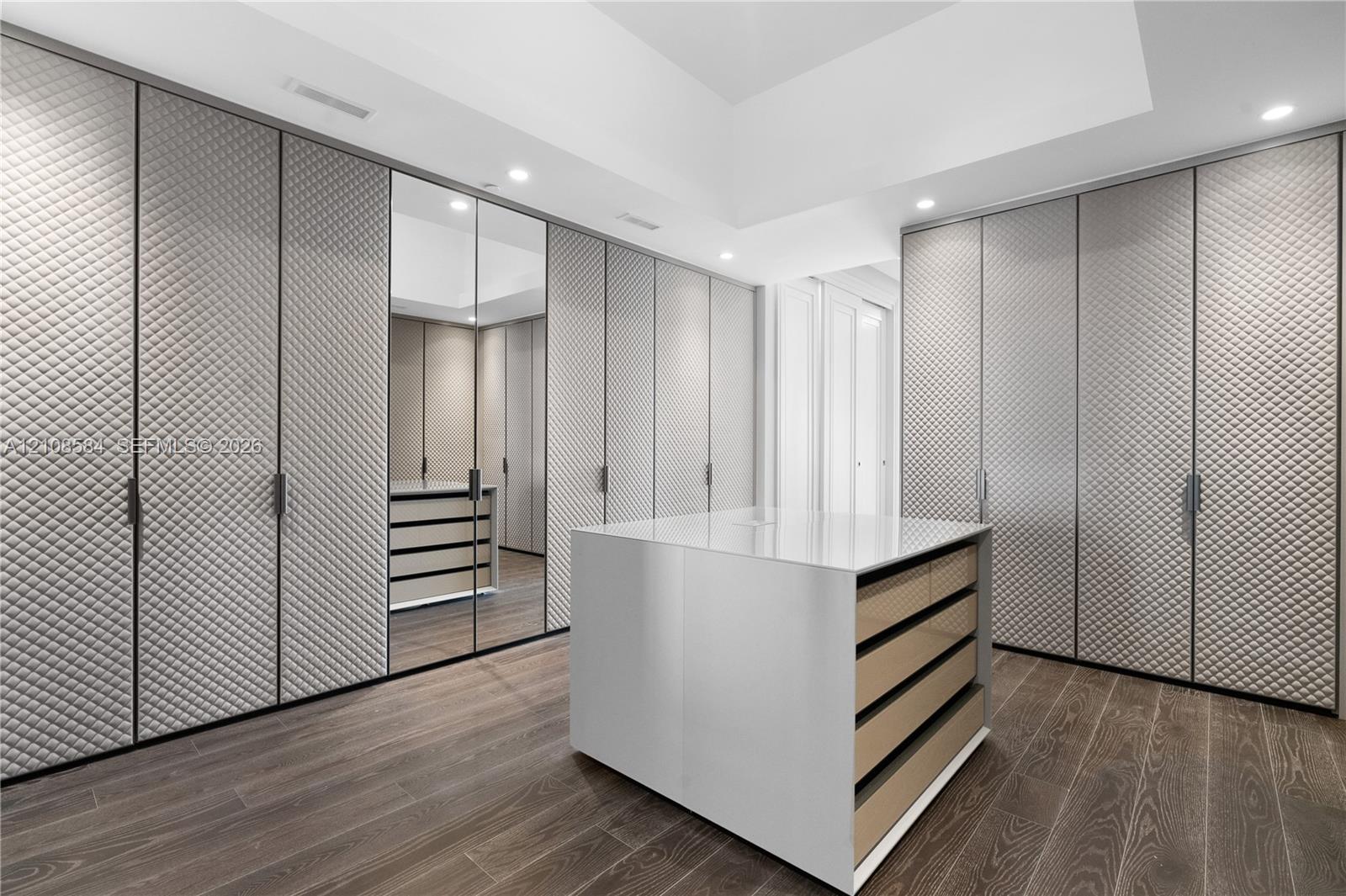 Floor-to-ceiling built-in cabinetry with diamond-quilted paneling, full-height mirrored wardrobe door, central island with smooth countertop, recessed lighting, and wood-finish flooring