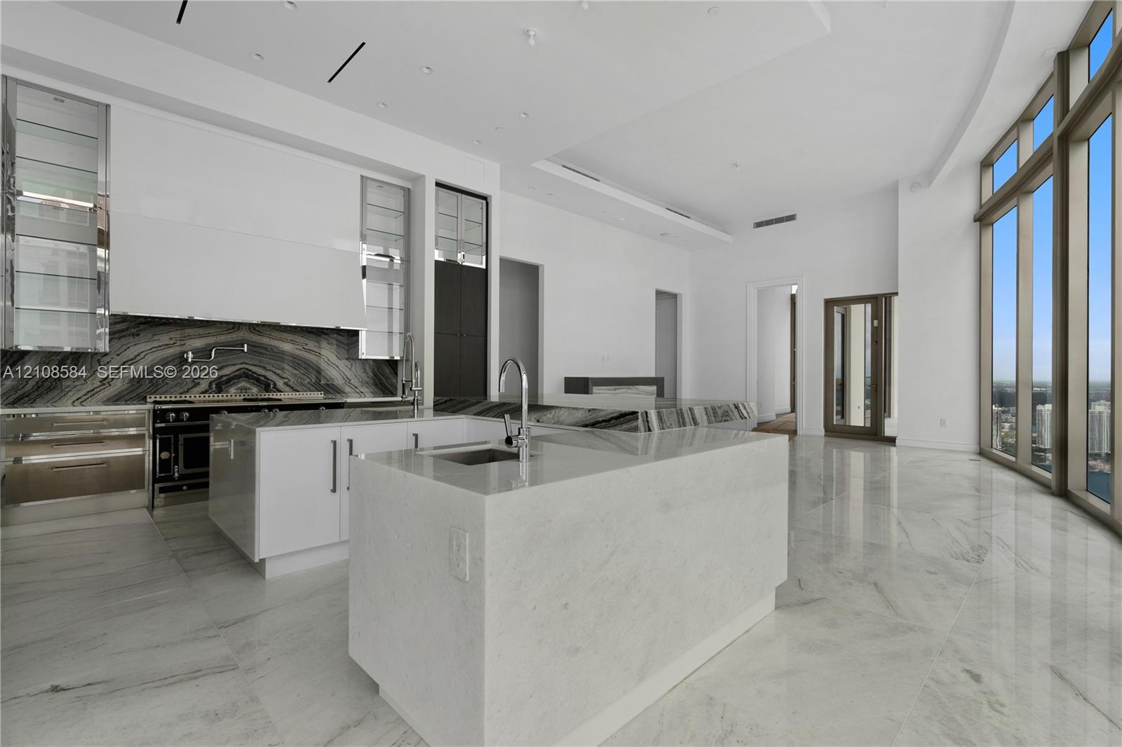 Contemporary kitchen featuring floor-to-ceiling windows, high-gloss cabinetry, stone-surfaced island with integrated sink, stone backsplash, and polished stone flooring