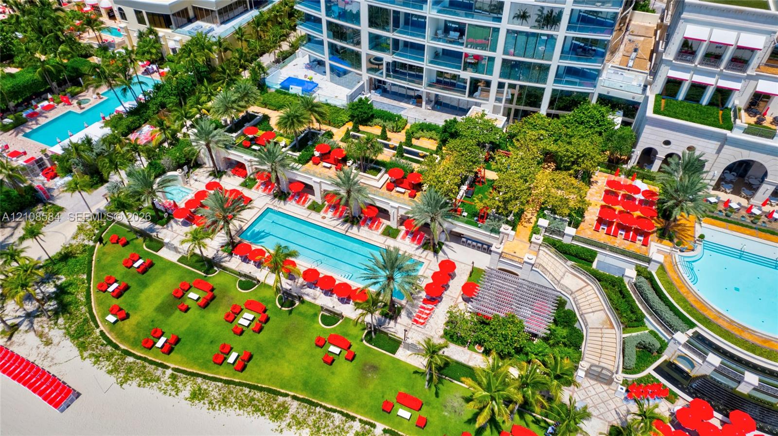 Aerial perspective of a coastal property featuring multiple swimming pools, tiered hardscaped sun decks, expansive manicured lawns, and integrated palm tree landscaping