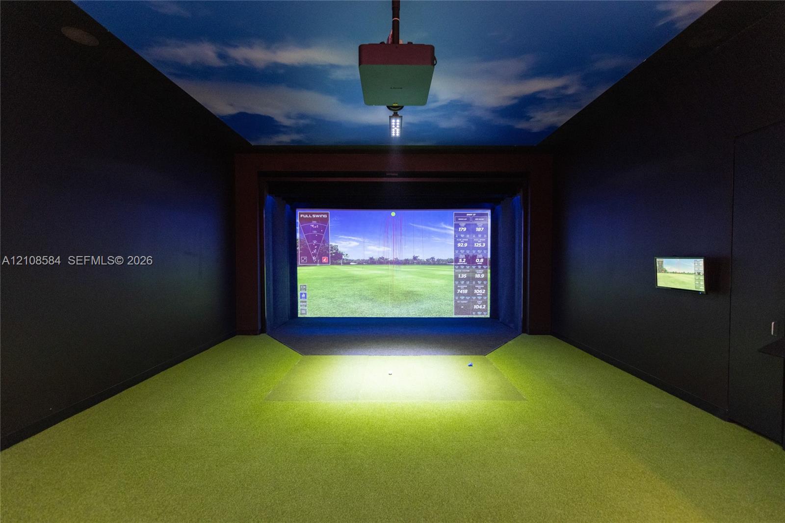 Dedicated simulation room featuring a projector system, integrated impact screen, wall-mounted display, and synthetic turf flooring with a painted sky ceiling motif