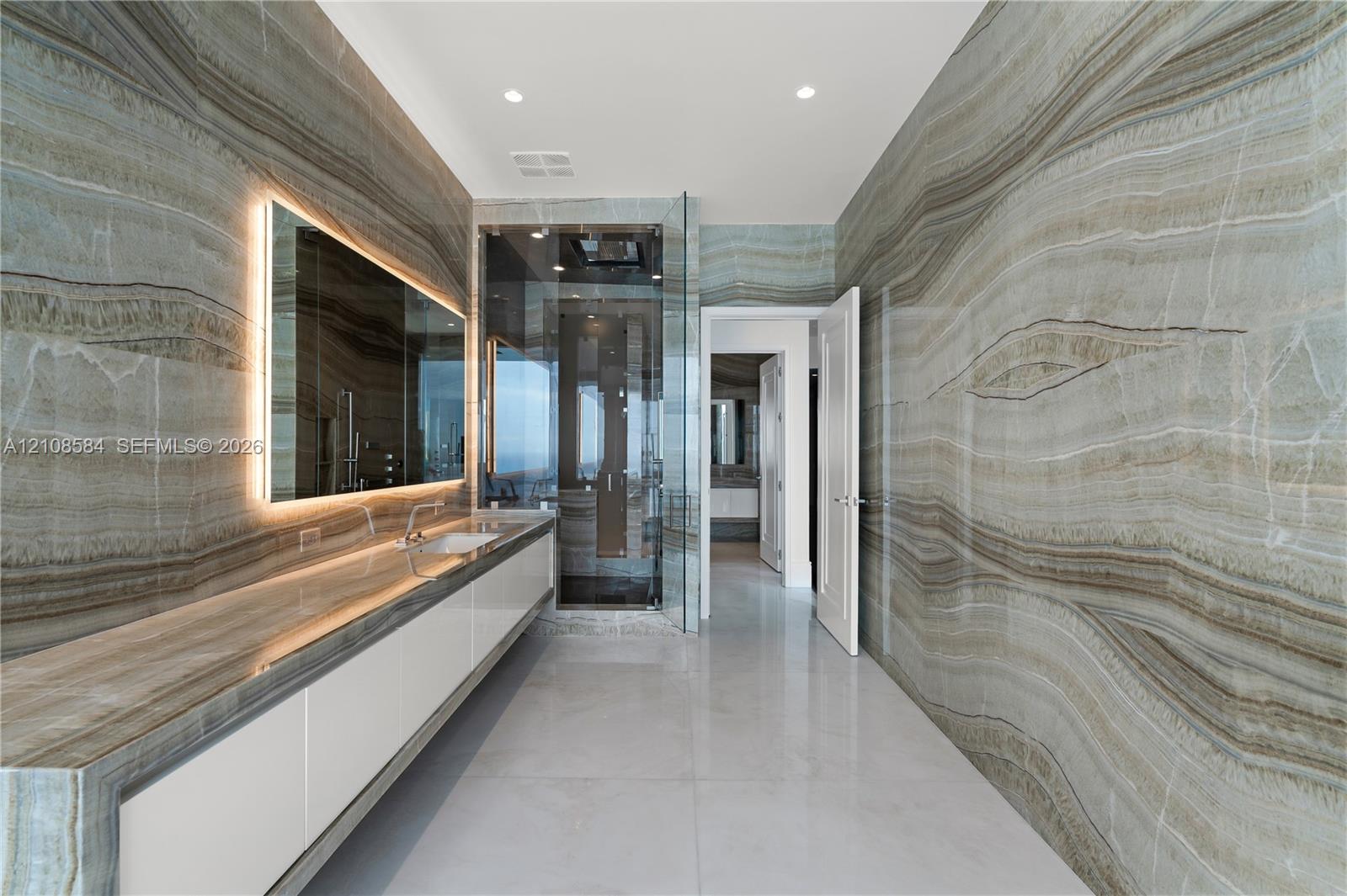 Full-height stone wall cladding, custom floating vanity with integrated stone countertop, LED-backlit mirror, large-format tile flooring, and glass-enclosed shower with rainfall showerhead