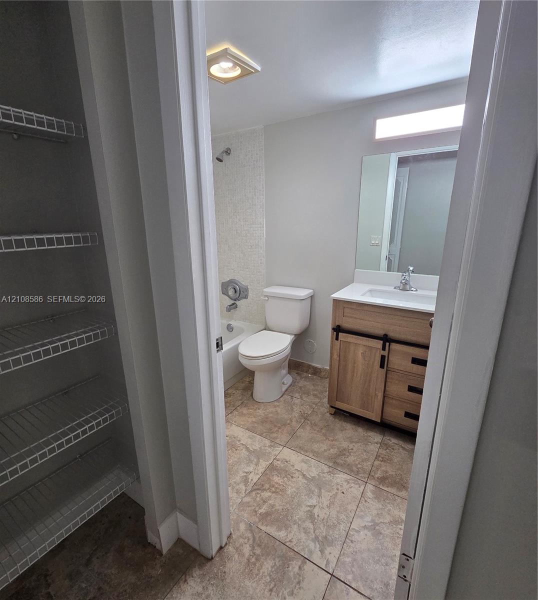 Bathroom featuring a wood-finish vanity with a white countertop, large wall-mounted mirror, tile flooring, bathtub with a tiled shower surround, and a walk-in storage area with wire shelving