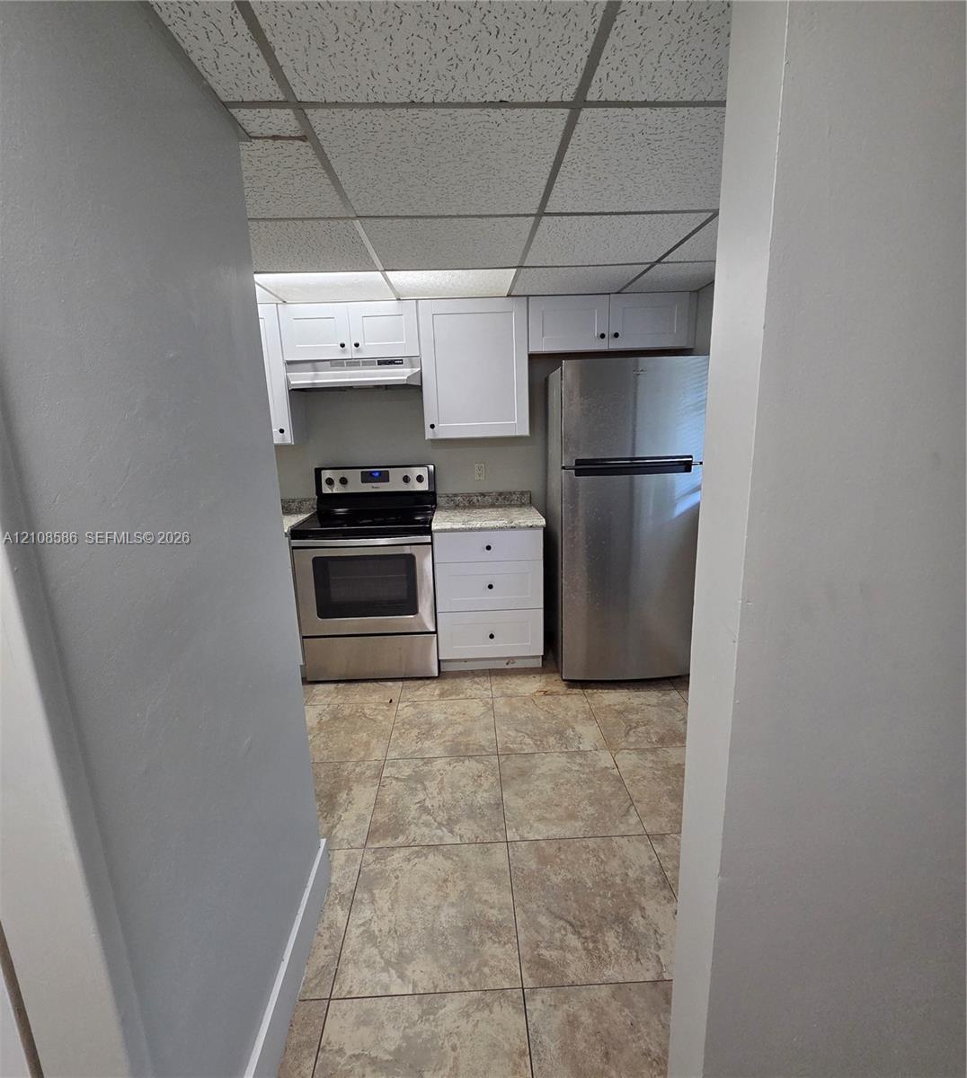 Kitchen featuring white shaker-style cabinetry, stainless steel appliances, speckled stone-finish countertops, tile flooring, and drop ceiling panels