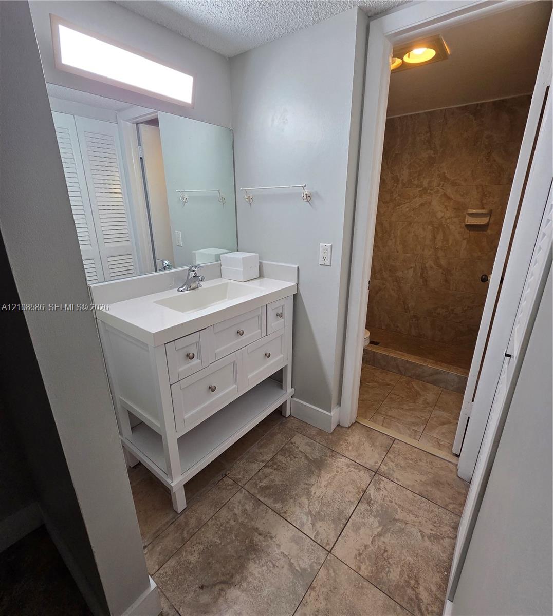 White vanity with integrated countertop and sink, large rectangular wall mirror, overhead linear lighting, and tile flooring with matching wall-tiled shower enclosure