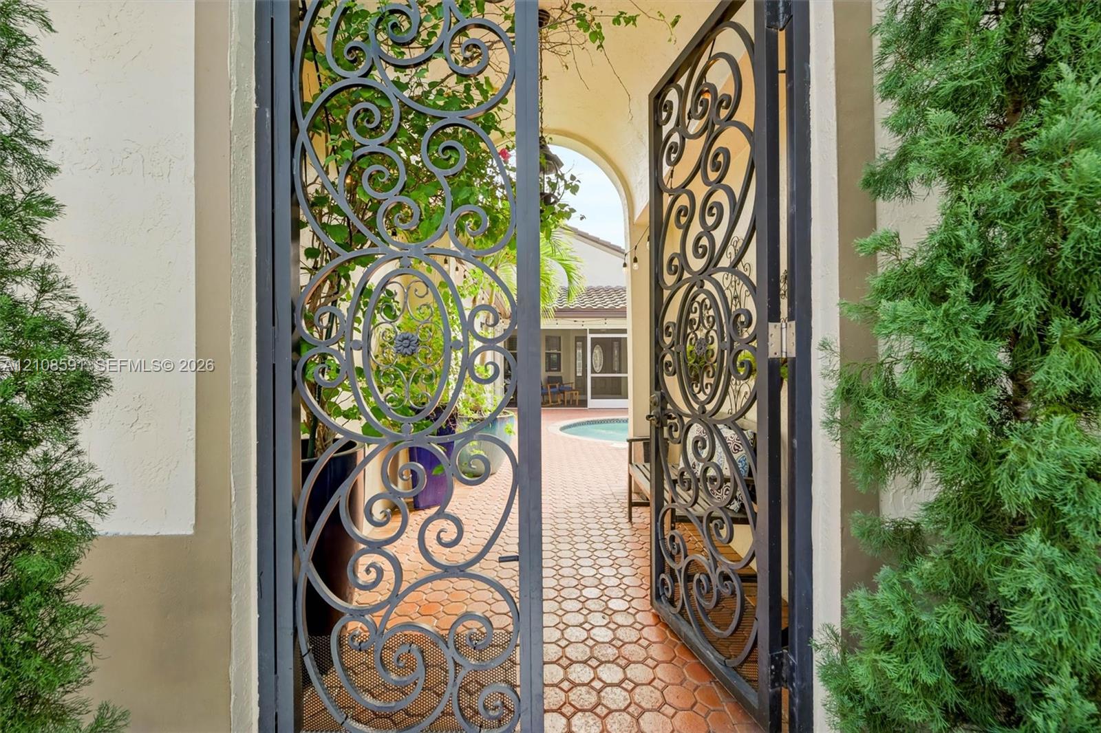 Decorative wrought-iron entry gate featuring intricate scrollwork, opening to a paved courtyard with a pool area