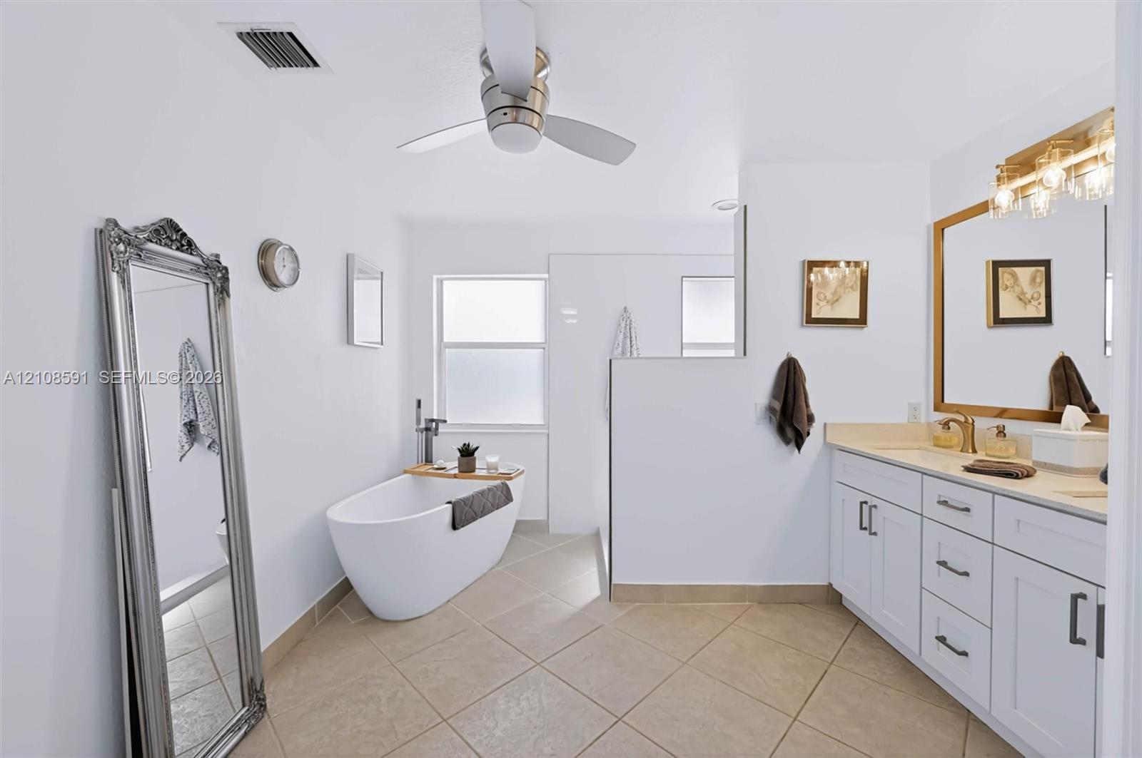 Bathroom featuring a freestanding soaking tub, white shaker-style cabinetry with neutral solid-surface countertops, tile flooring, and a multi-light vanity fixture