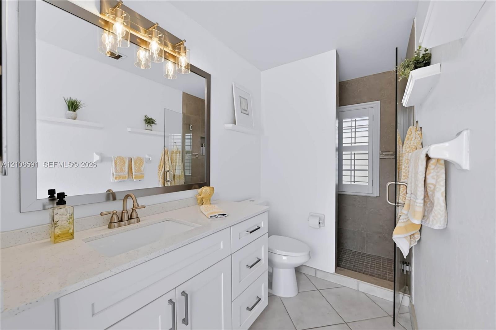 Bathroom vanity featuring white shaker-style cabinetry, a light-toned solid surface countertop, an integrated rectangular sink, a multi-light fixture, and a walk-in shower with a glass door and neutral wall tile