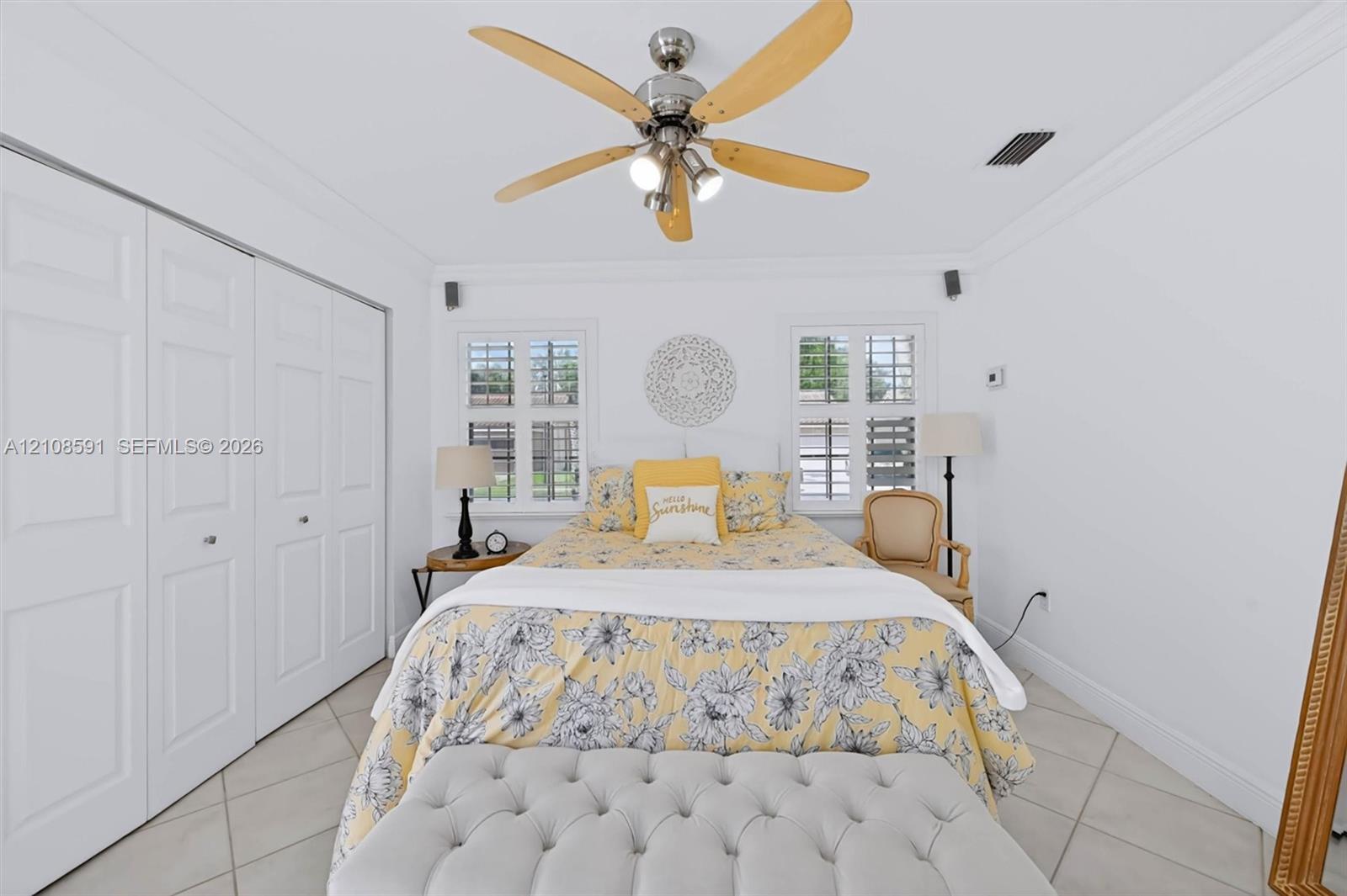 Neutral-toned tile flooring, ceiling fan with integrated lighting, dual windows with plantation shutters, and floor-to-ceiling closets with paneled doors