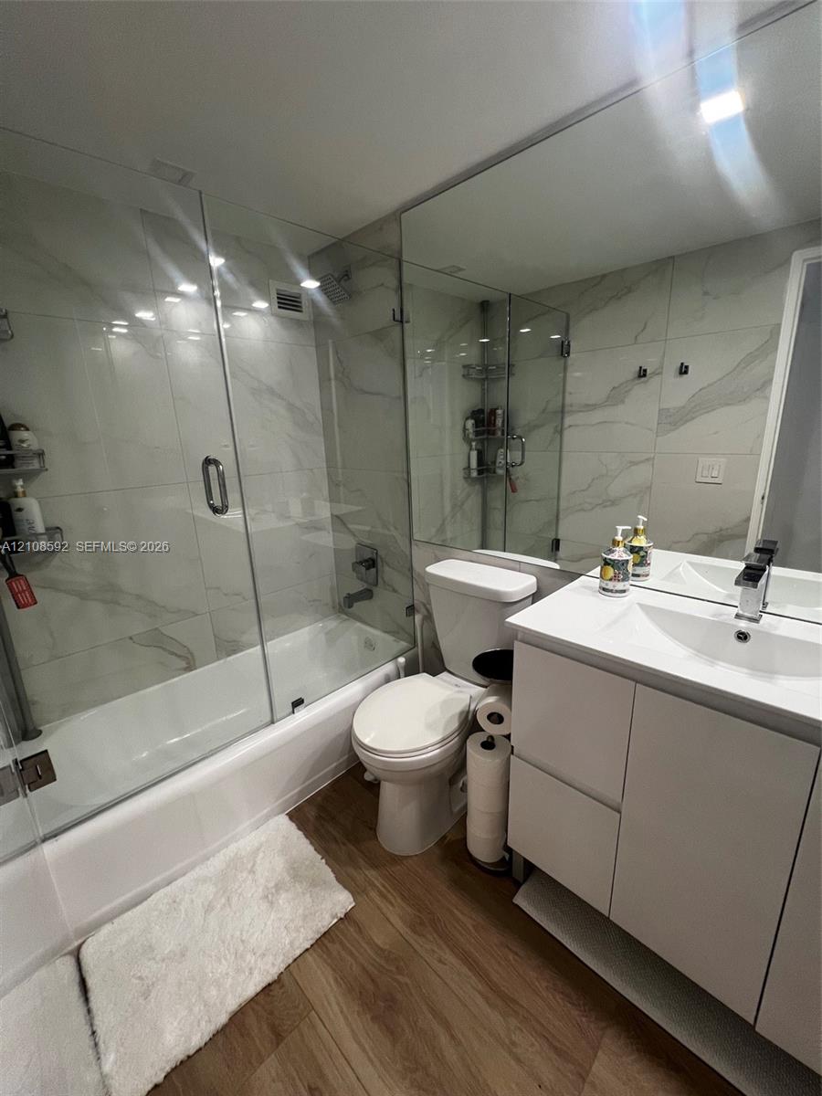 Bathroom featuring a vanity with a white countertop and rectangular basin, full-width wall-mounted mirror, wood-finish flooring, and a glass-enclosed shower with large-format wall tiling