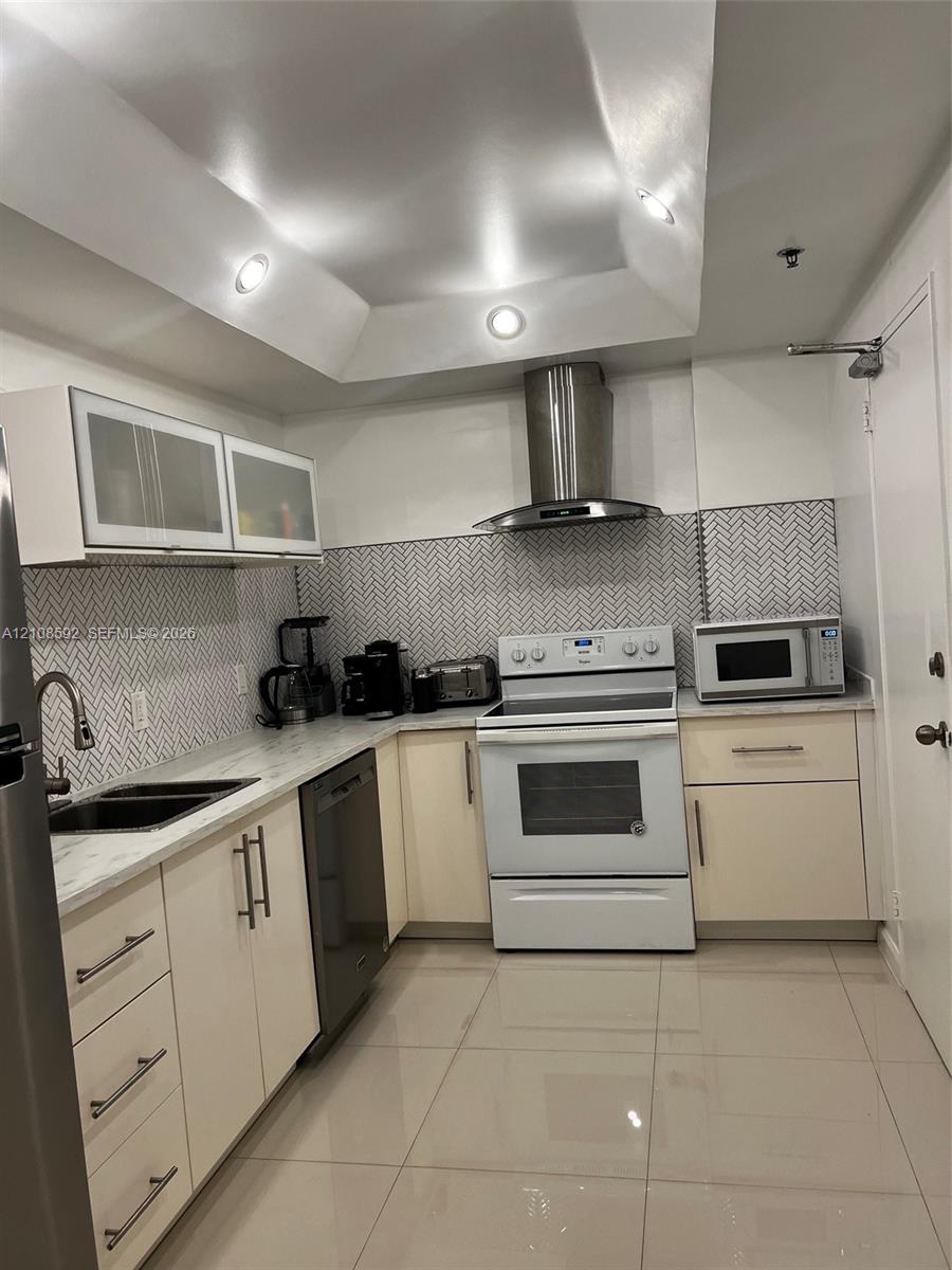 Kitchen featuring high-gloss cabinetry, stone-finish countertops, herringbone-patterned tile backsplash, stainless steel chimney-style range hood, and recessed tray ceiling lighting