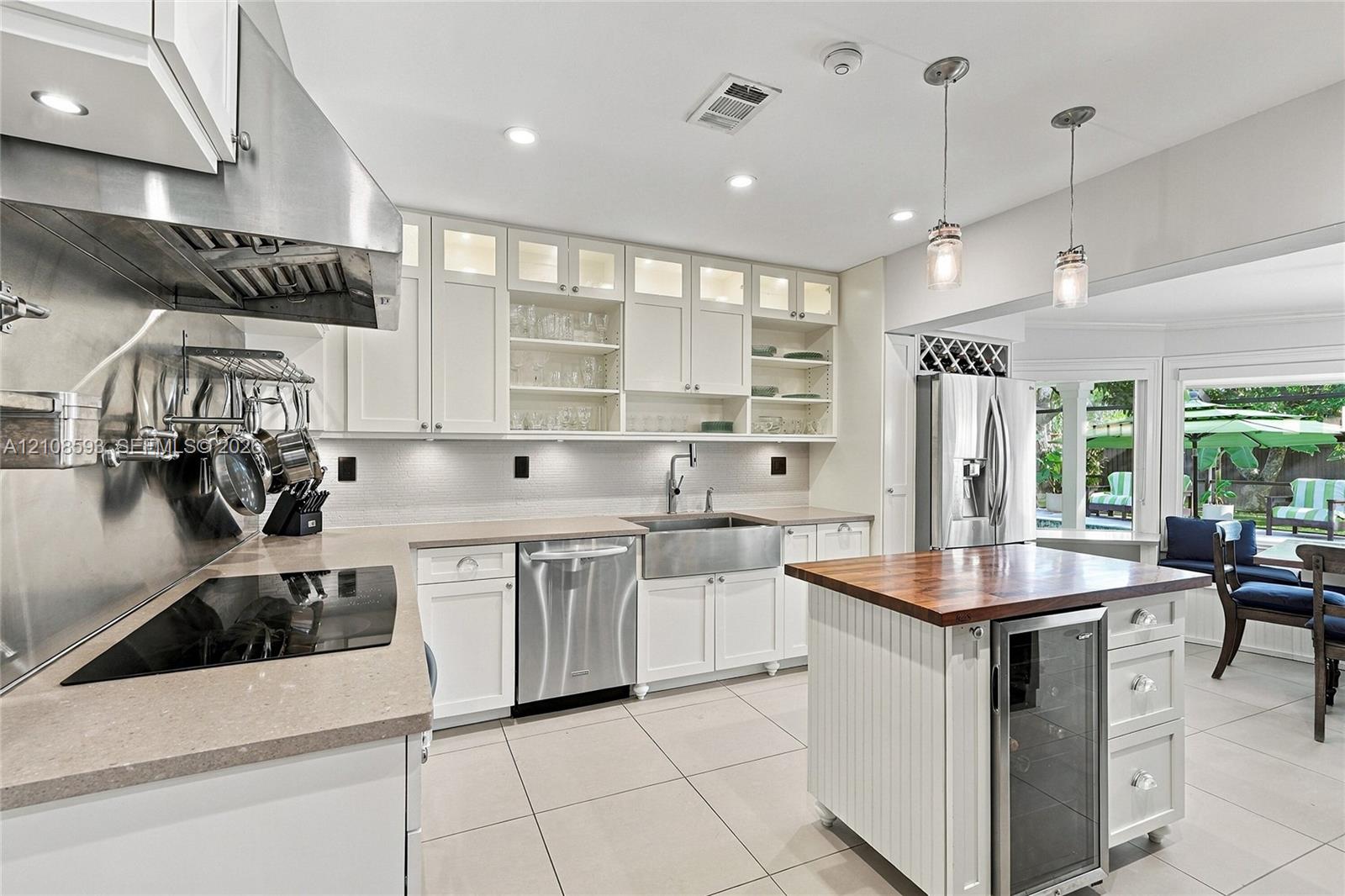 Kitchen featuring white cabinetry with glass-front uppers, stainless steel farmhouse sink, professional-grade range hood, butcher-block topped island with built-in wine refrigeration, and stone-finish countertops