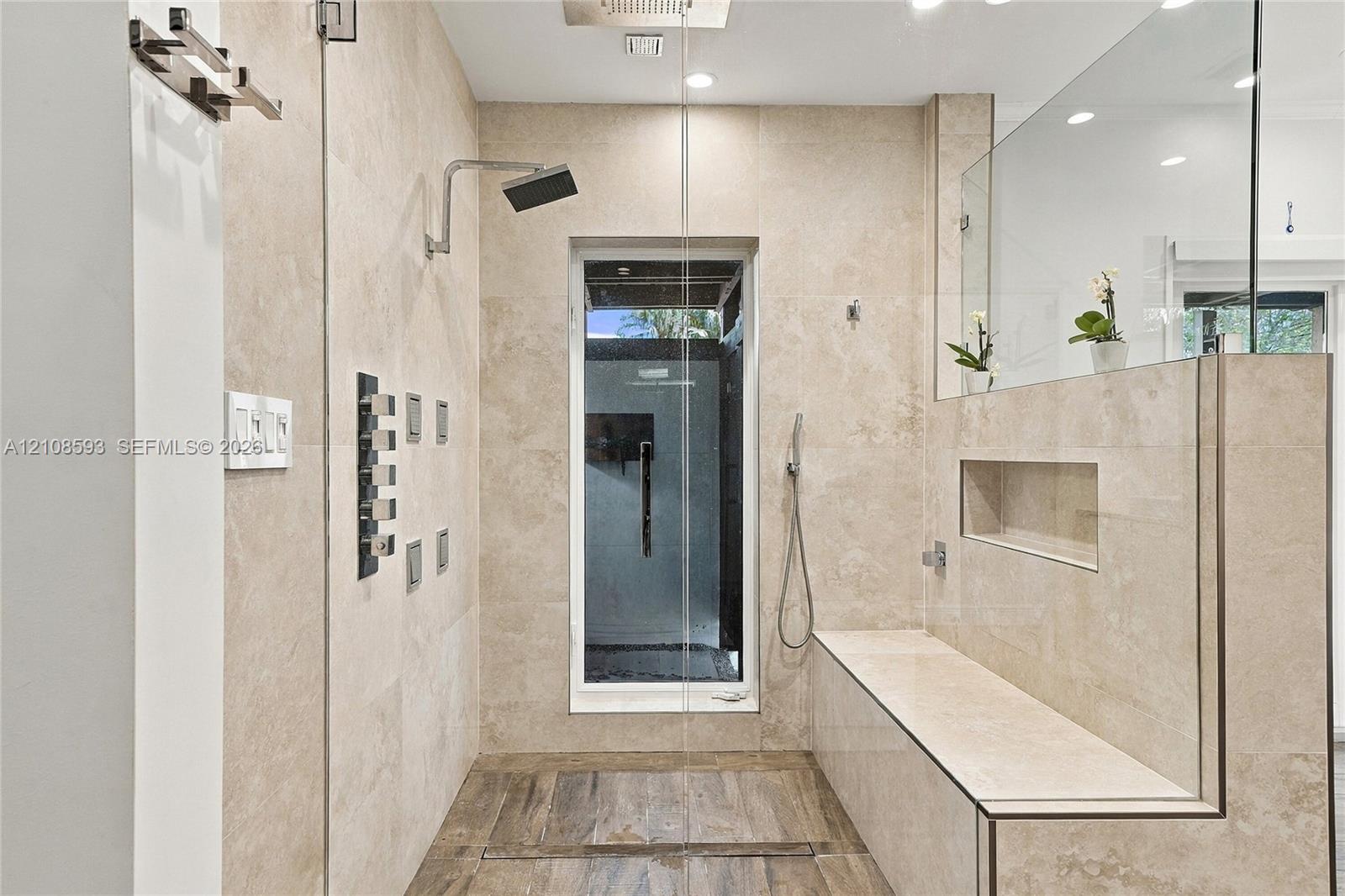 Walk-in shower featuring large-format neutral stone wall tile, multi-head shower system with a rain shower head, built-in stone bench with integrated recessed shelving, and wood-finish flooring