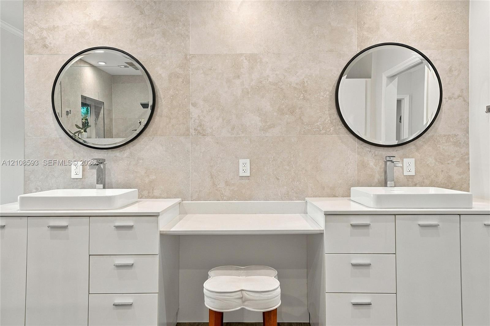 Dual white cabinetry with white stone countertops, integrated center vanity station, dual vessel sinks, wall-mounted round mirrors with matte black frames, and neutral tile wall surround