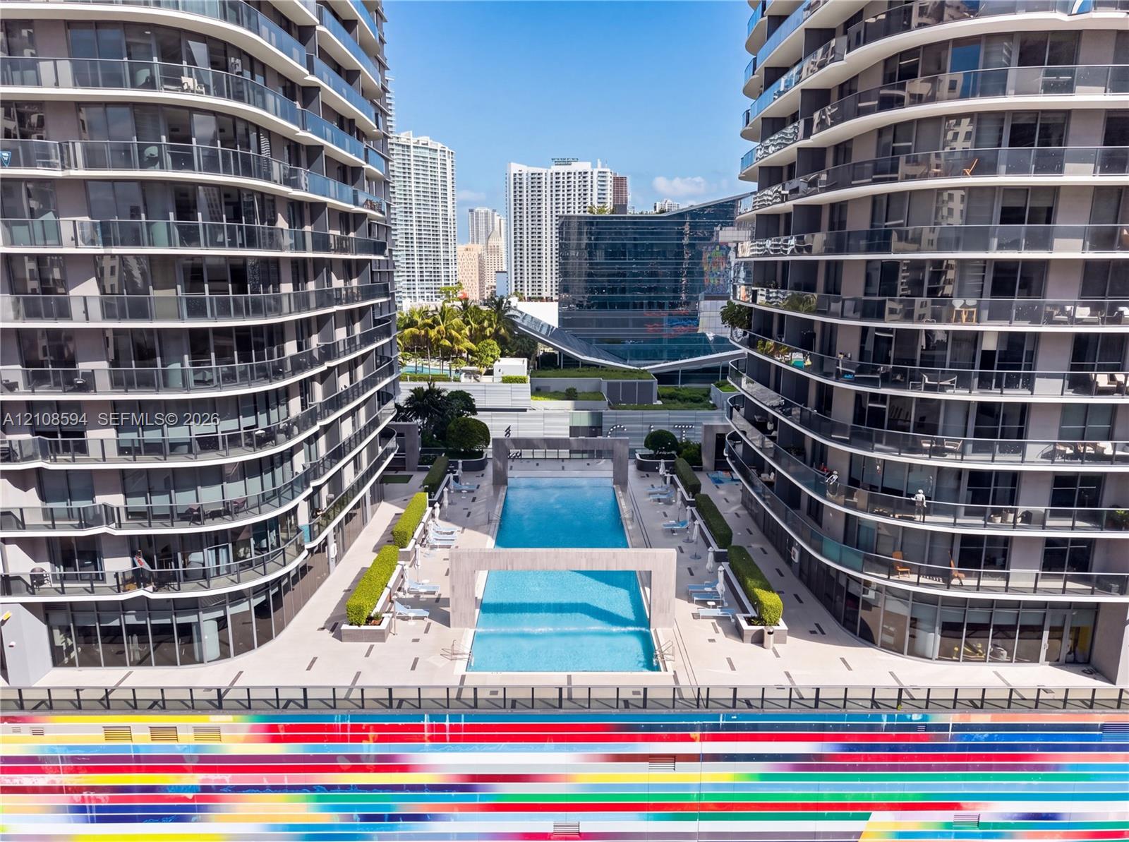 Outdoor pool featuring concrete deck surfaces, decorative stone archways, manicured hedges, and multi-story high-rise buildings with glass-paneled balconies