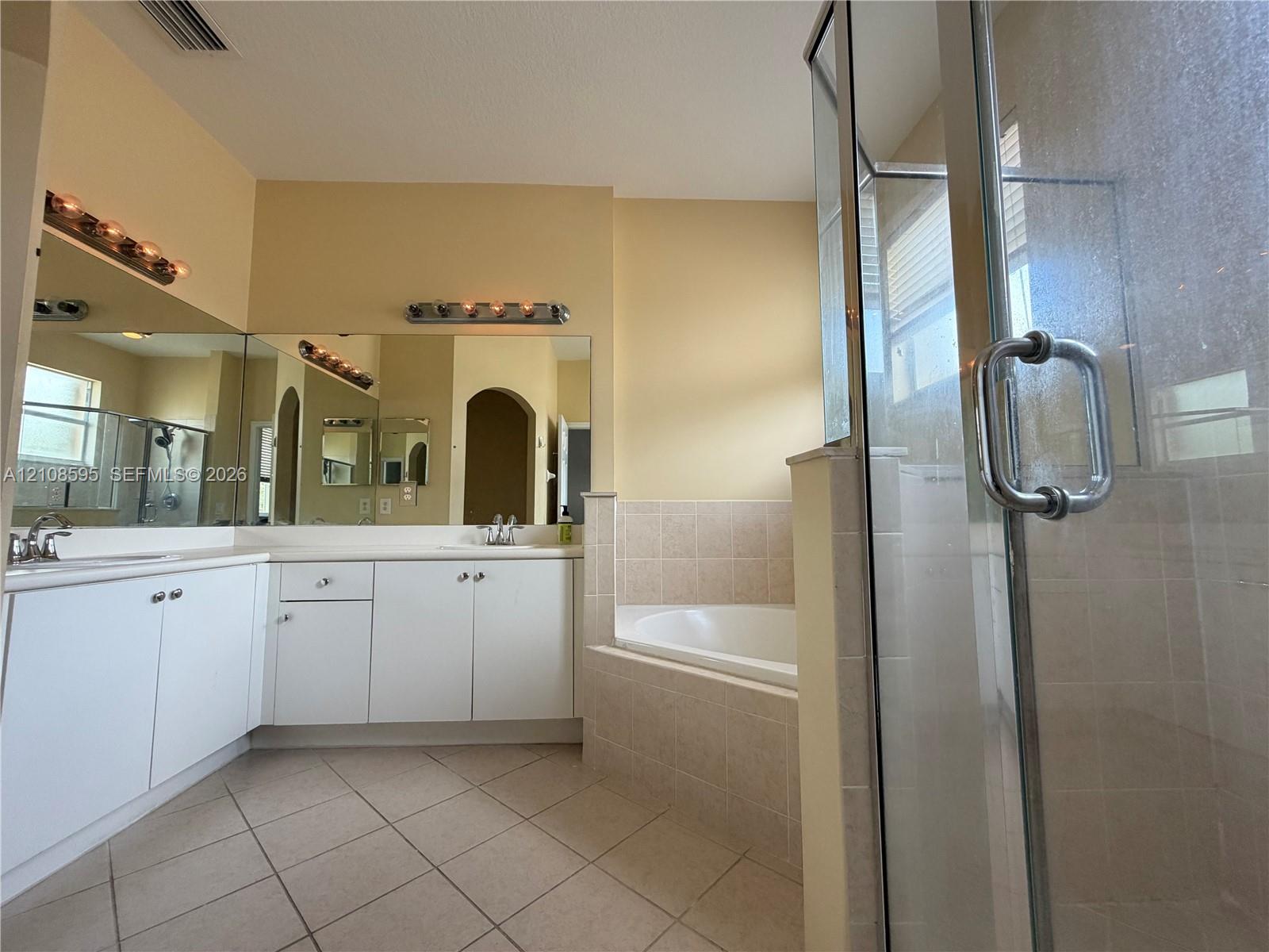 Bathroom featuring white cabinetry, dual sinks, tile-surround soaking tub, separate glass-enclosed shower, and tile flooring