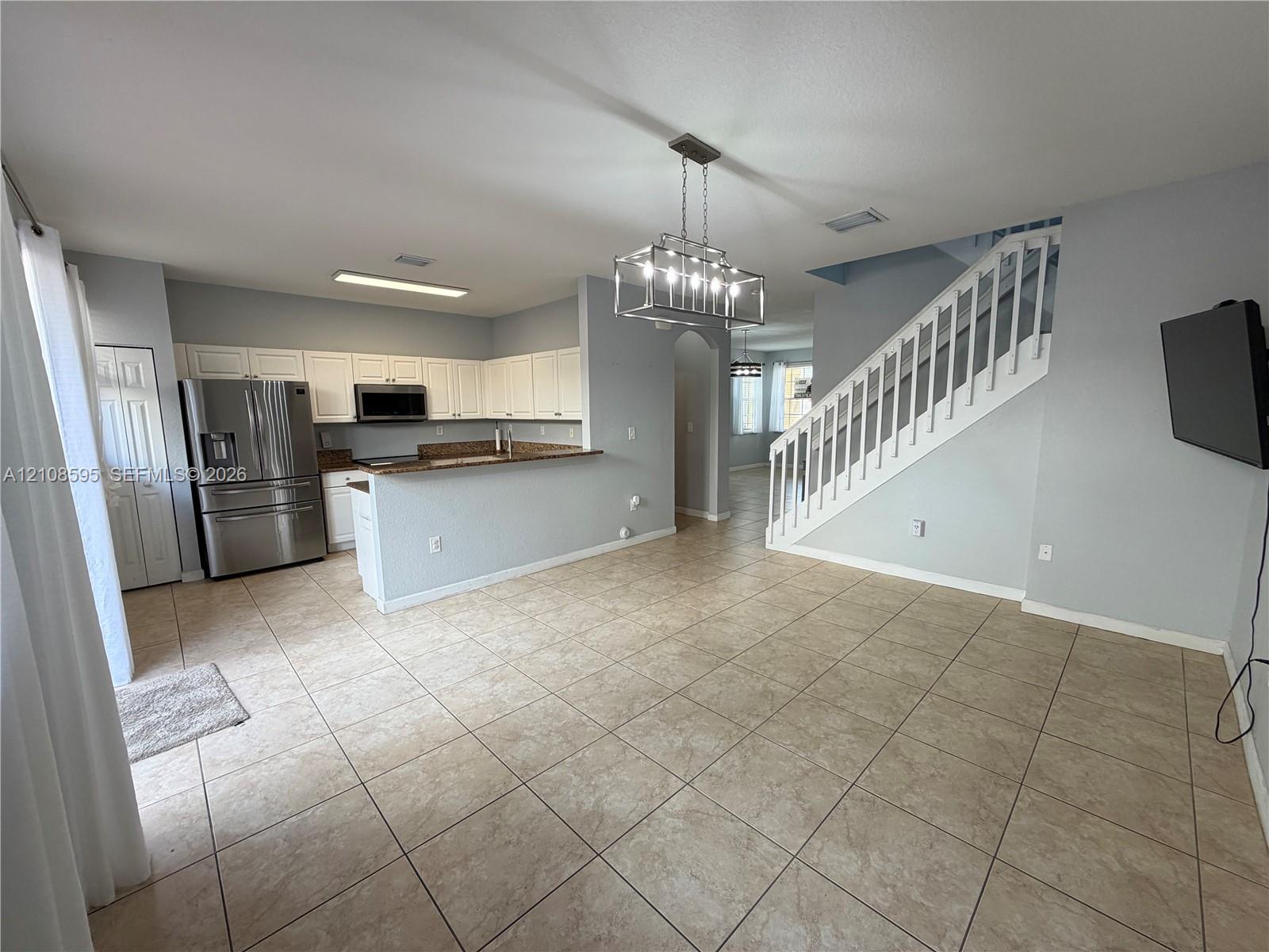 Open-concept space featuring ceramic tile flooring, kitchen with white cabinetry and stone countertops, stainless steel appliances, and a built-in staircase with white railings