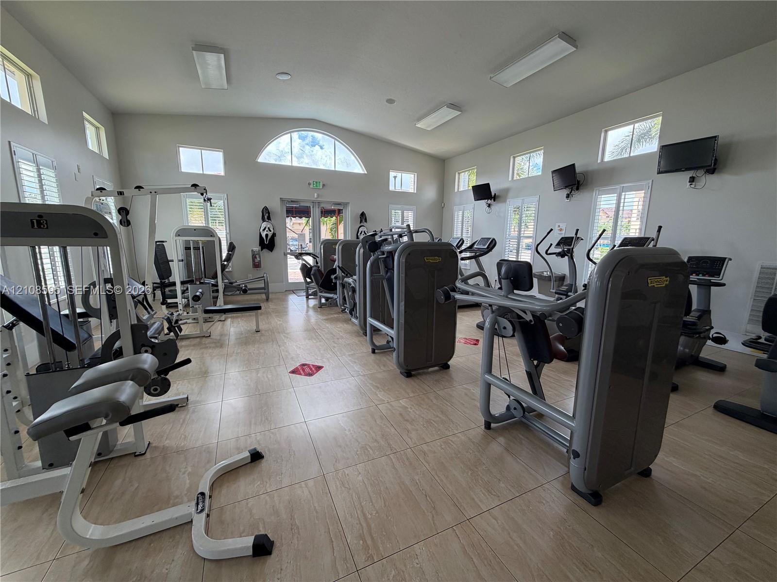 Fitness center featuring large-format tile flooring, vaulted ceilings with recessed lighting, and multiple windows including an arched architectural accent