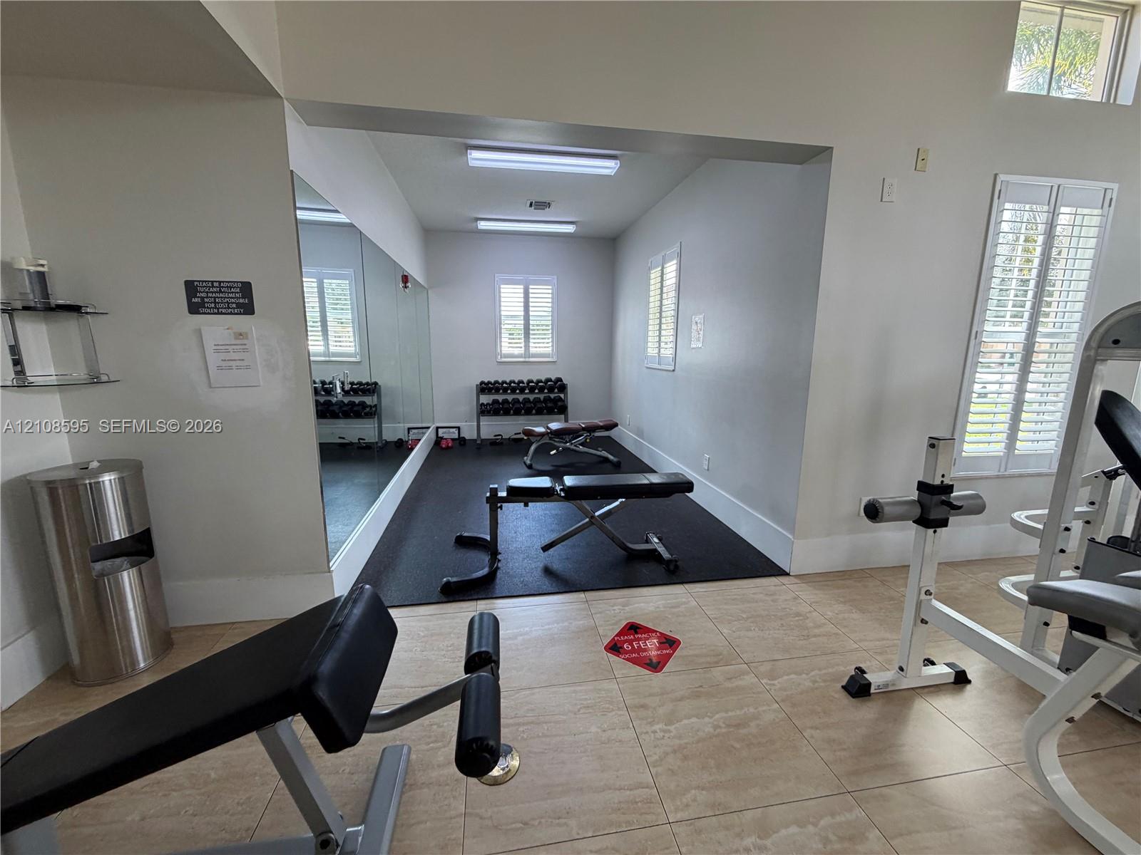 Fitness area featuring tile flooring, a rubber-floored section, full-length wall mirror, windows with plantation shutters, and overhead fluorescent lighting
