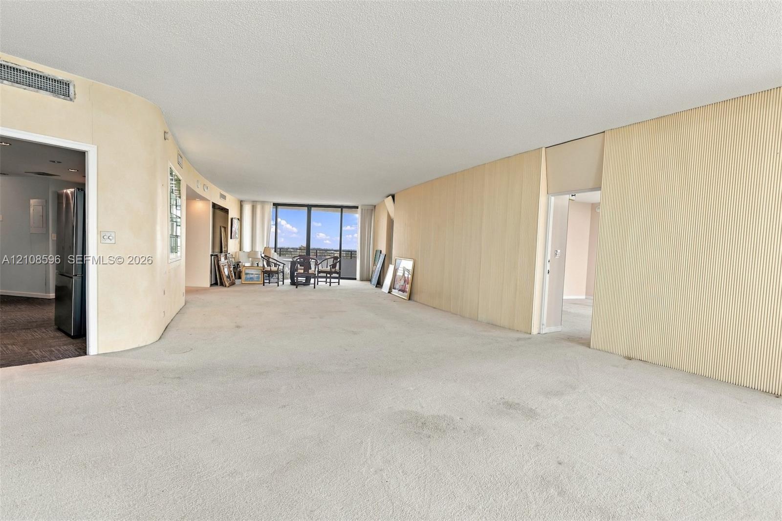 Open-concept floor plan featuring expansive light-toned wall paneling, curved wall architecture, floor-to-ceiling glass sliding doors, and neutral wall-to-wall carpeting