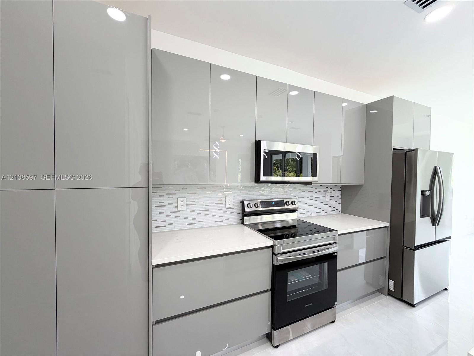 Modern kitchen featuring high-gloss gray cabinetry, white solid-surface countertops, a mosaic tile backsplash, and stainless steel appliances