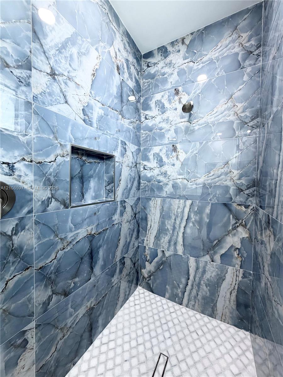 Shower stall featuring full-height blue-toned stone-patterned wall tiles, integrated recessed shelving, white geometric mosaic floor tiling, and a metallic shower fixture