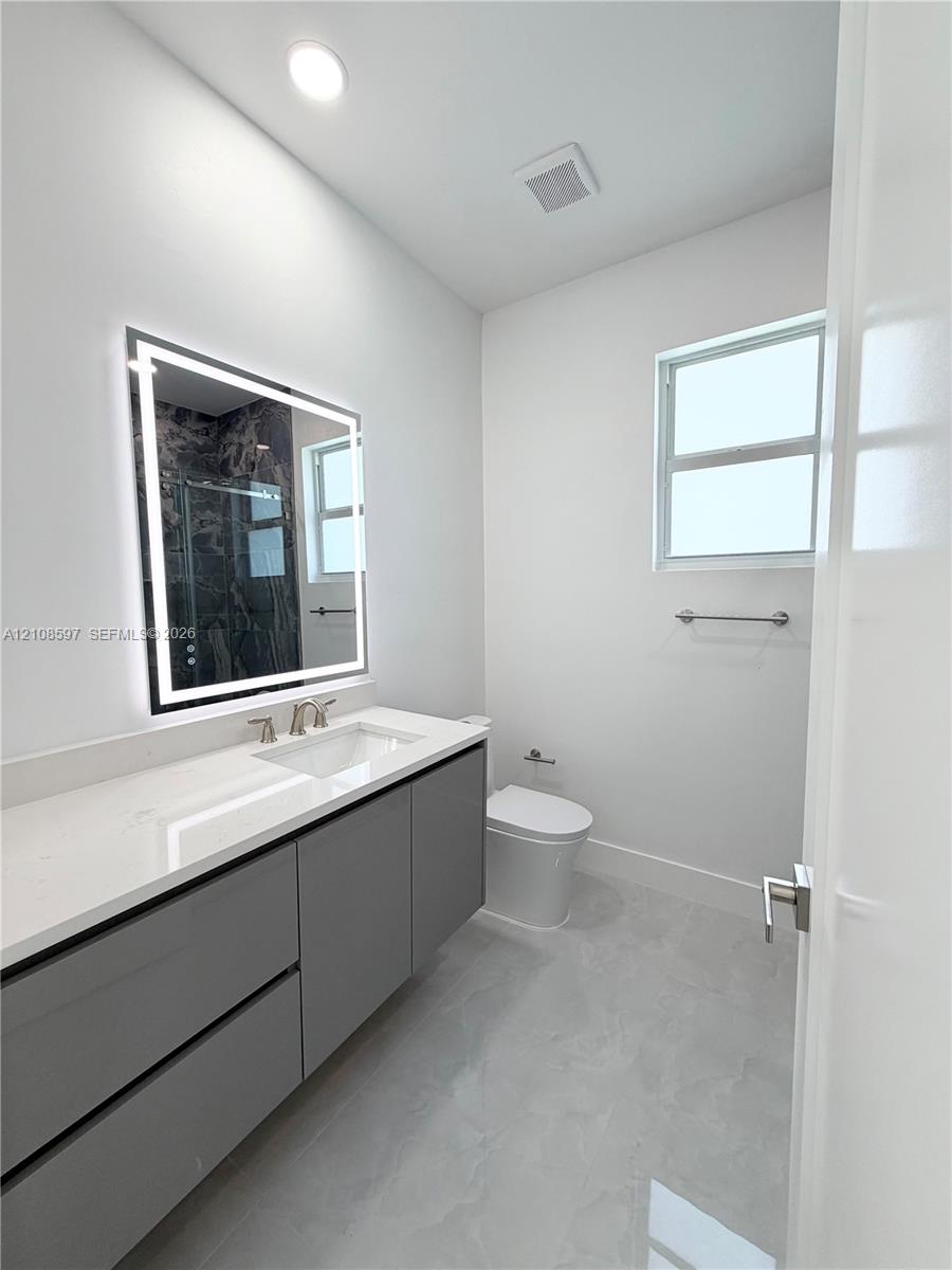 Modern vanity featuring a stone countertop, integrated sink, and illuminated rectangular mirror, complemented by grey cabinetry, light-toned flooring, and a window for natural light