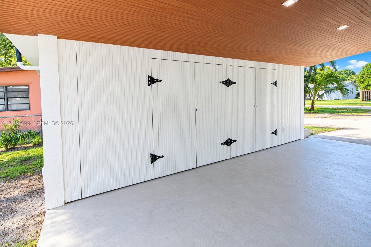 Covered carport featuring a finished wood-tone ceiling with recessed lighting, smooth concrete flooring, and white vertical-paneled storage cabinetry with decorative black iron hardware