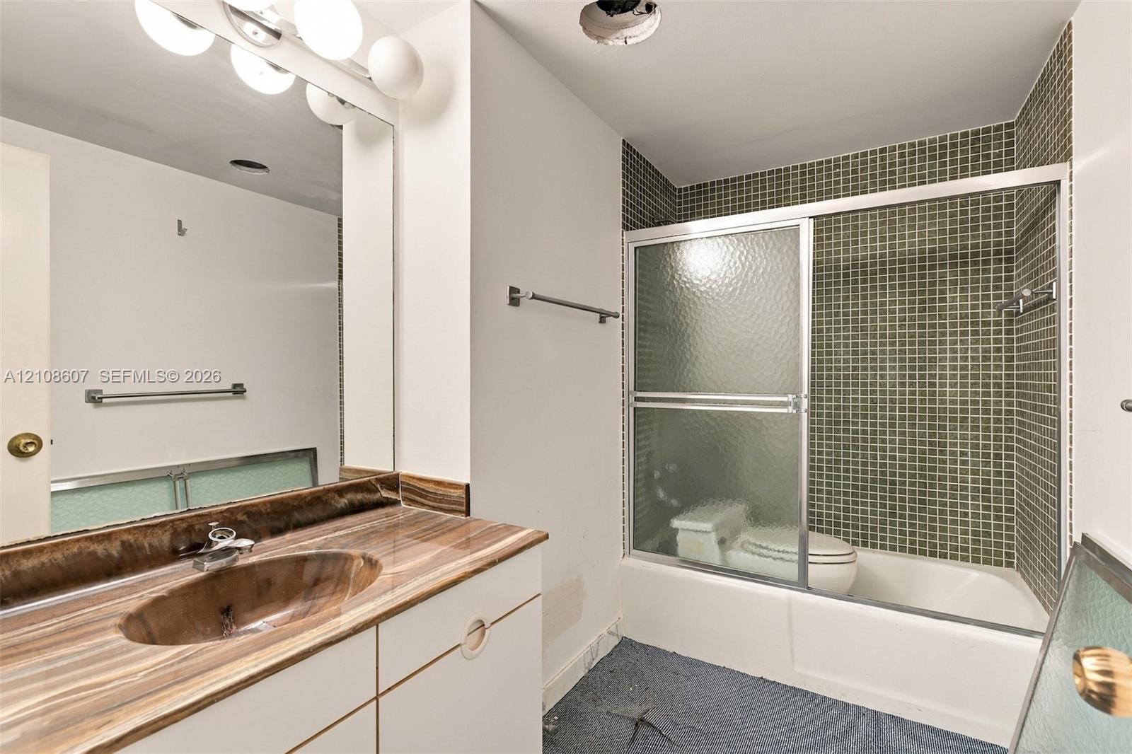 Vanity featuring a natural stone-patterned countertop with a recessed basin, integrated wall-width mirror, shower and tub combination with glass sliding doors, and green mosaic tile surround