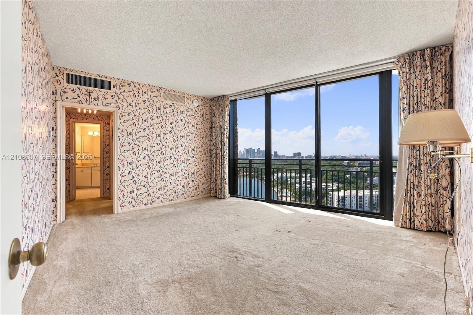 Floor-to-ceiling windows featuring panoramic city and water scenery, black metal balcony railing, and neutral wall-to-wall carpeting