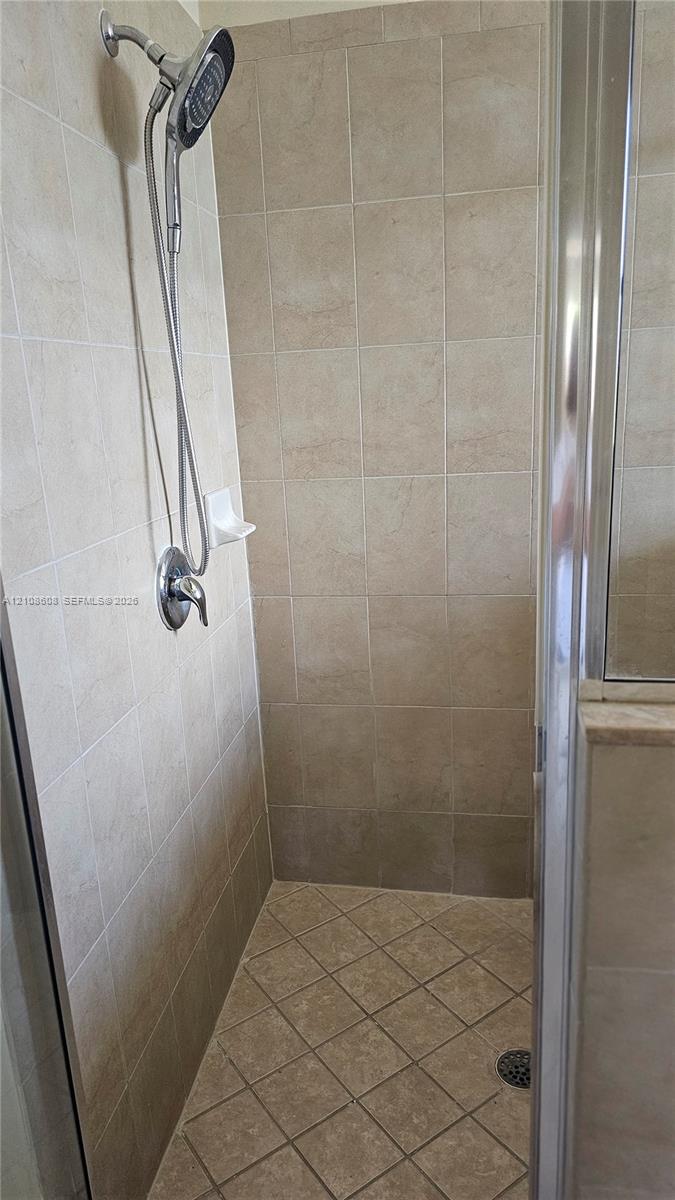 Shower stall featuring neutral-toned wall tiling, diamond-patterned floor tile, a handheld shower fixture, and a glass-enclosed door
