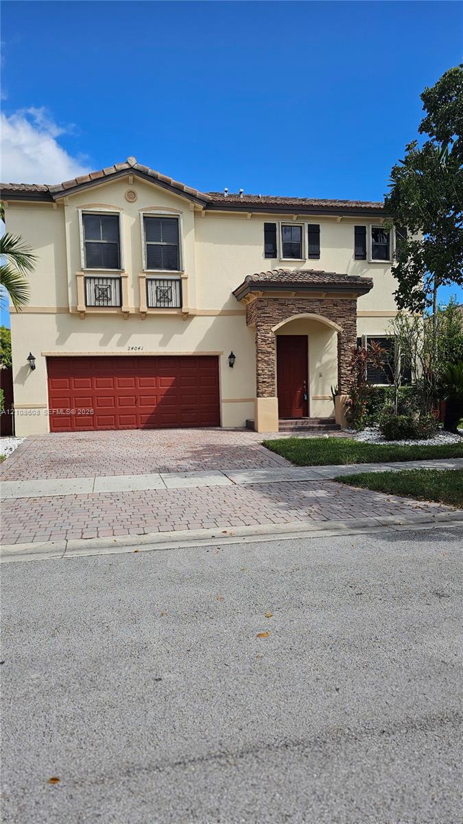 Two-story exterior featuring a stone-veneer entryway, paver driveway, tiled roof, decorative balcony railings, and a multi-panel garage door