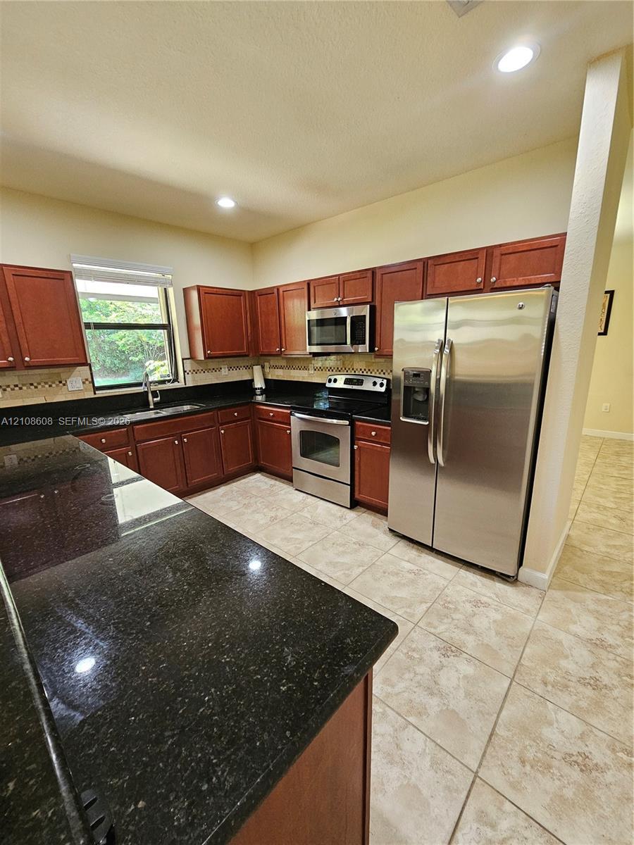 Kitchen featuring stained-wood cabinetry, dark stone countertops, stainless steel appliances, ceramic tile flooring, and recessed lighting