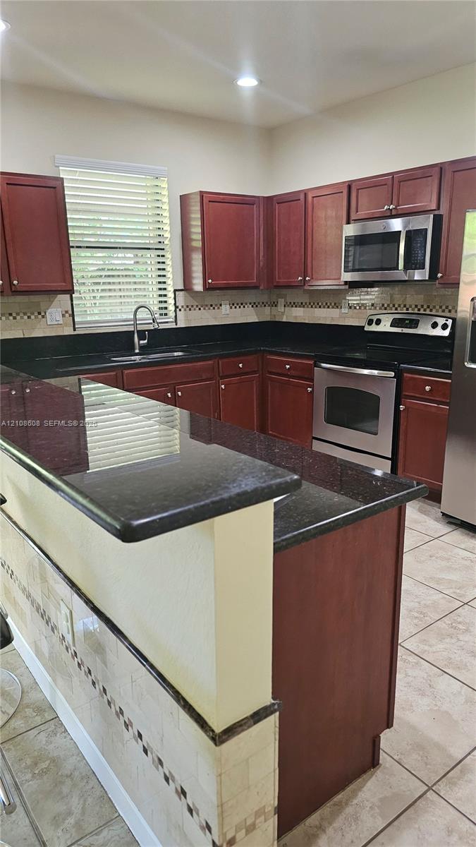 Kitchen featuring dark wood cabinetry, contrasting stone countertops, stainless steel appliances, ceramic tile flooring, and a stone tile backsplash