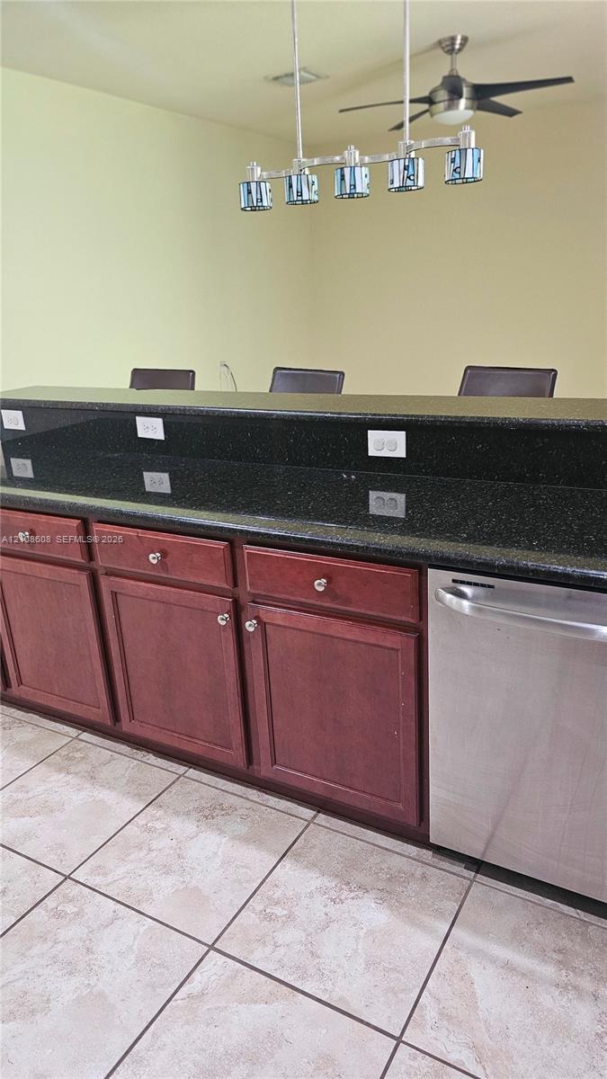 Kitchen island featuring a dark stone countertop, stained wood cabinetry, stainless steel dishwasher, five-light pendant fixture, and ceramic tile flooring