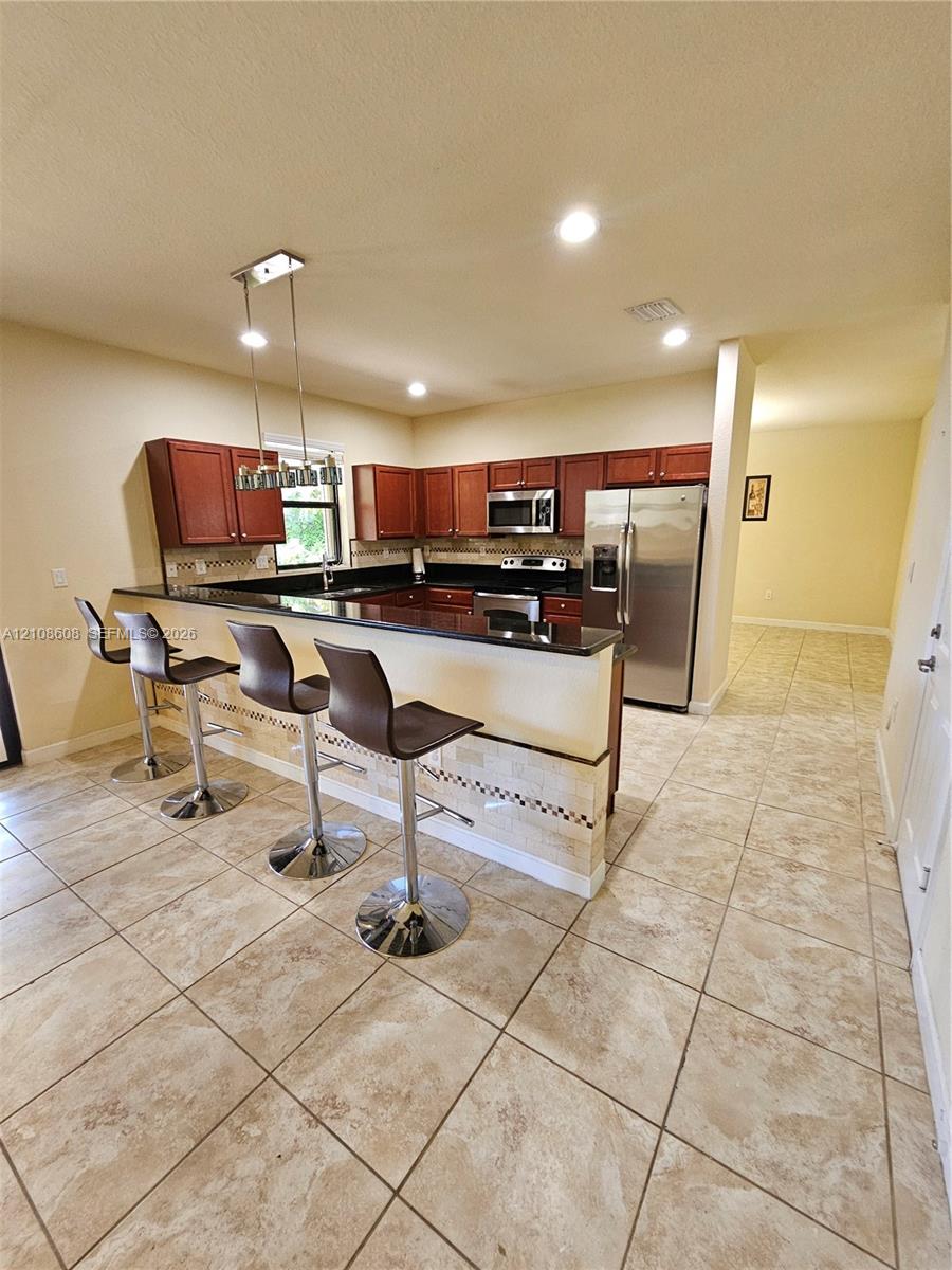 Kitchen featuring wood cabinetry, dark stone countertops, stainless steel appliances, ceramic tile flooring, and a stone-tiled breakfast bar